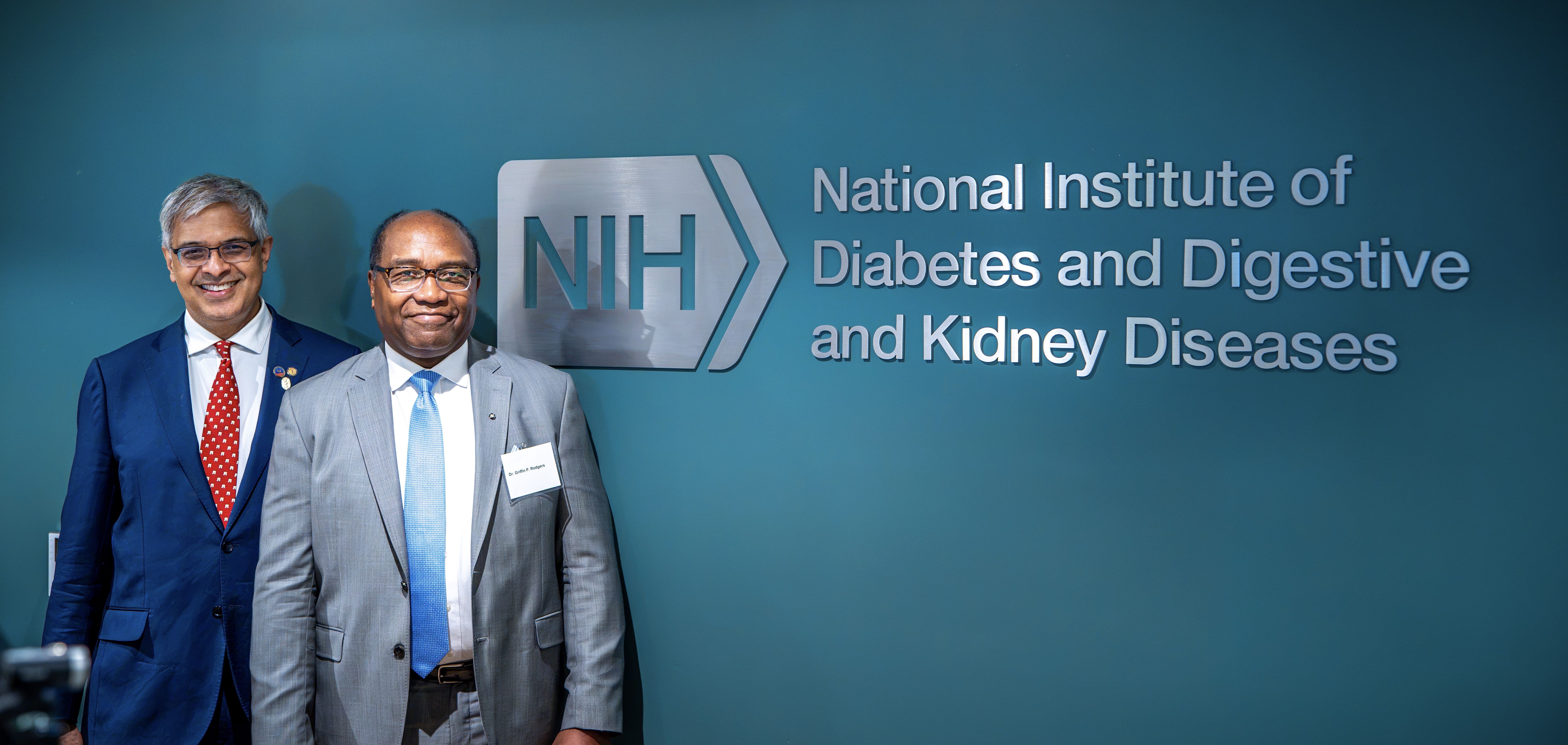 Two men in suits stand smiling in front of a teal wall with the NIH logo and the sign National Institute Of Diabetes and Digestive and Kidney Diseases.