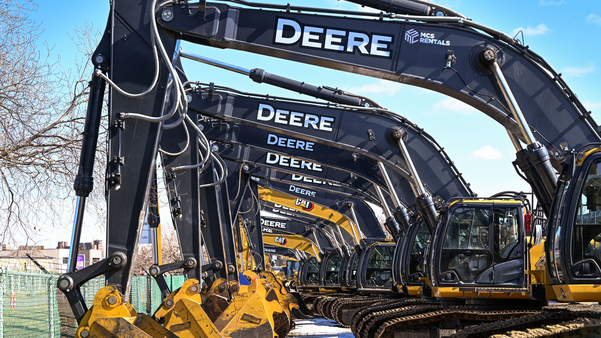 Row of Deere excavators with black arms and yellow buckets, parked side by side under a blue sky, near a green fence and leafless trees.