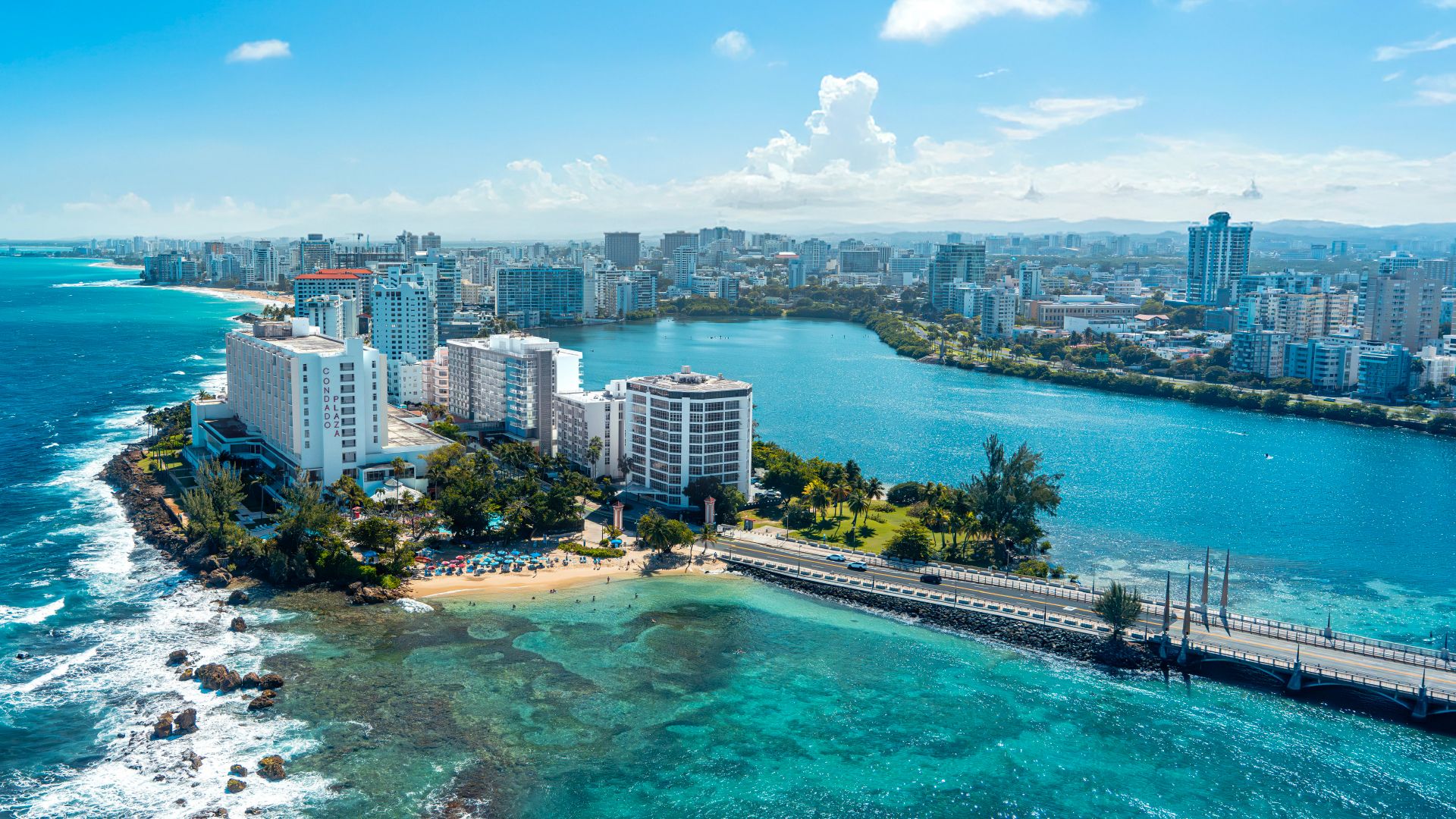 Aerial view of a sunny coastal city with a turquoise bay, a sandy beach, a bridge, and a cluster of high-rise buildings along the shoreline; clear blue sky with fluffy clouds.