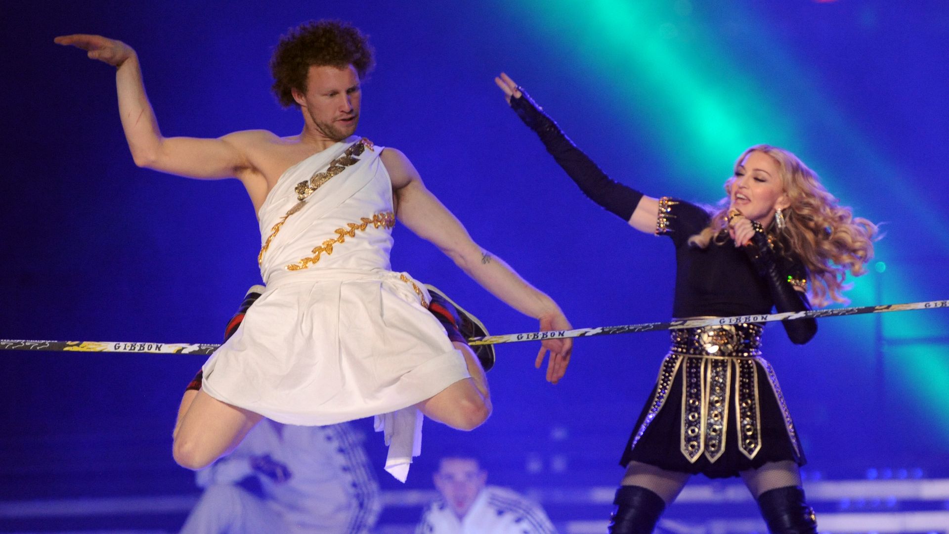 A man dances on a slackline while wearing a toga while MAdonna performs next to him
