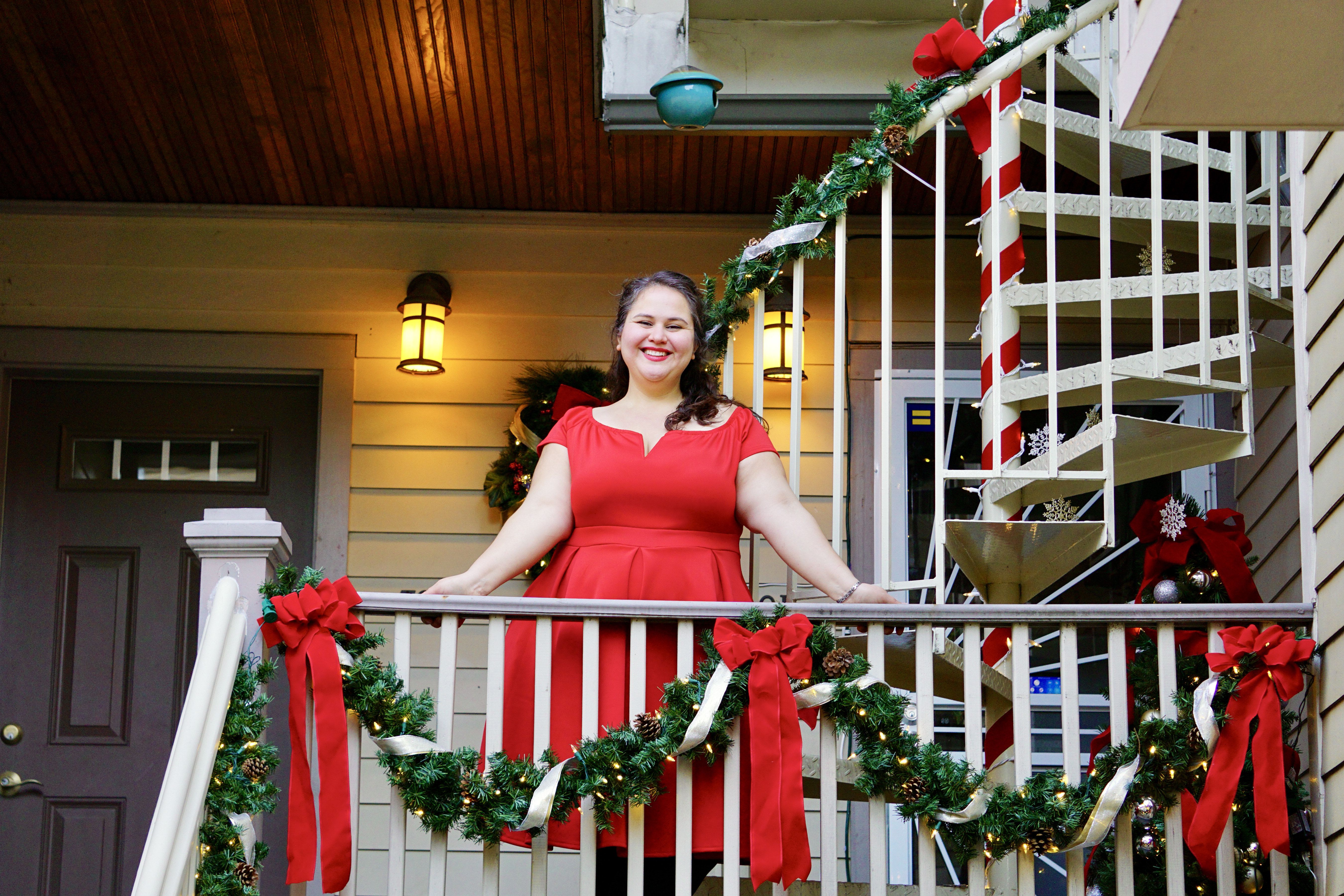 Smiling woman in a red dress stands on a porch decorated with Christmas garlands, red bows, pine cones, and white ribbon, with warm yellow lights and a candy-cane striped spiral staircase.