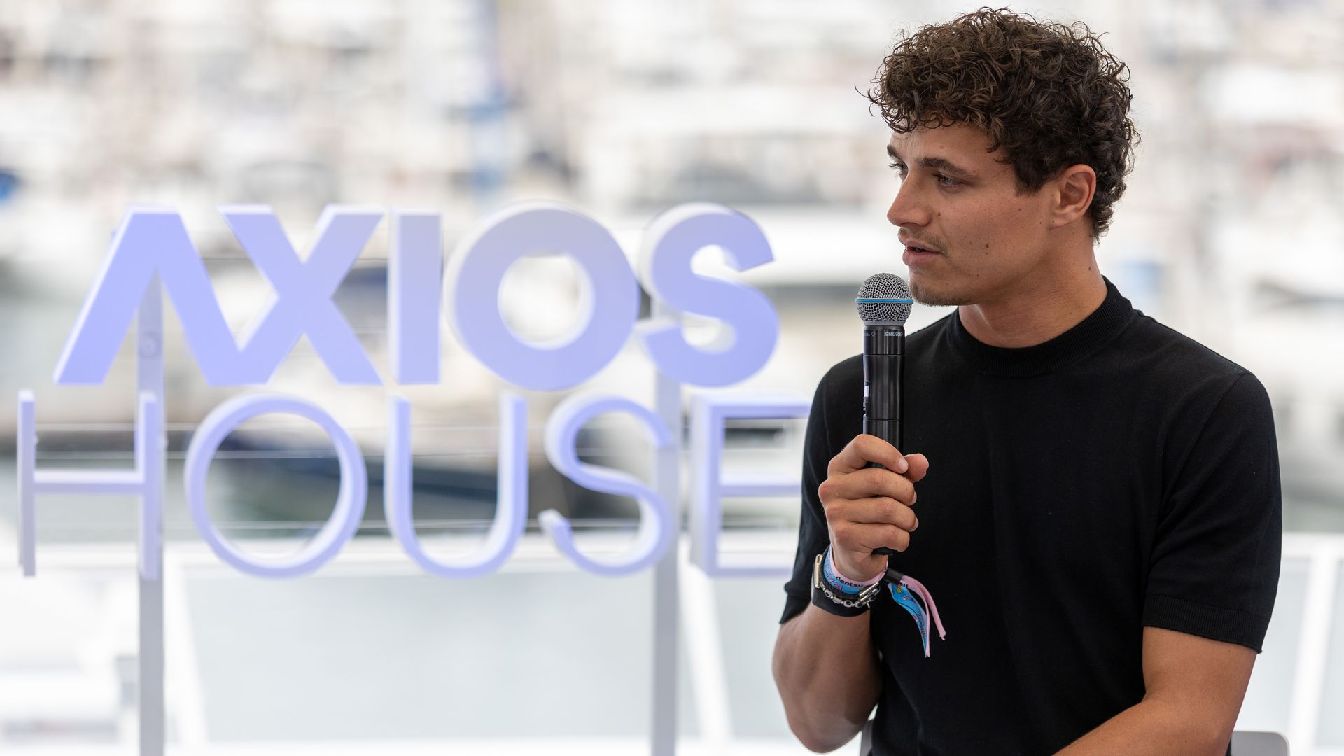 Young man with curly hair in a black shirt holds a microphone, standing beside large lavender letters with a blurred harbor backdrop.