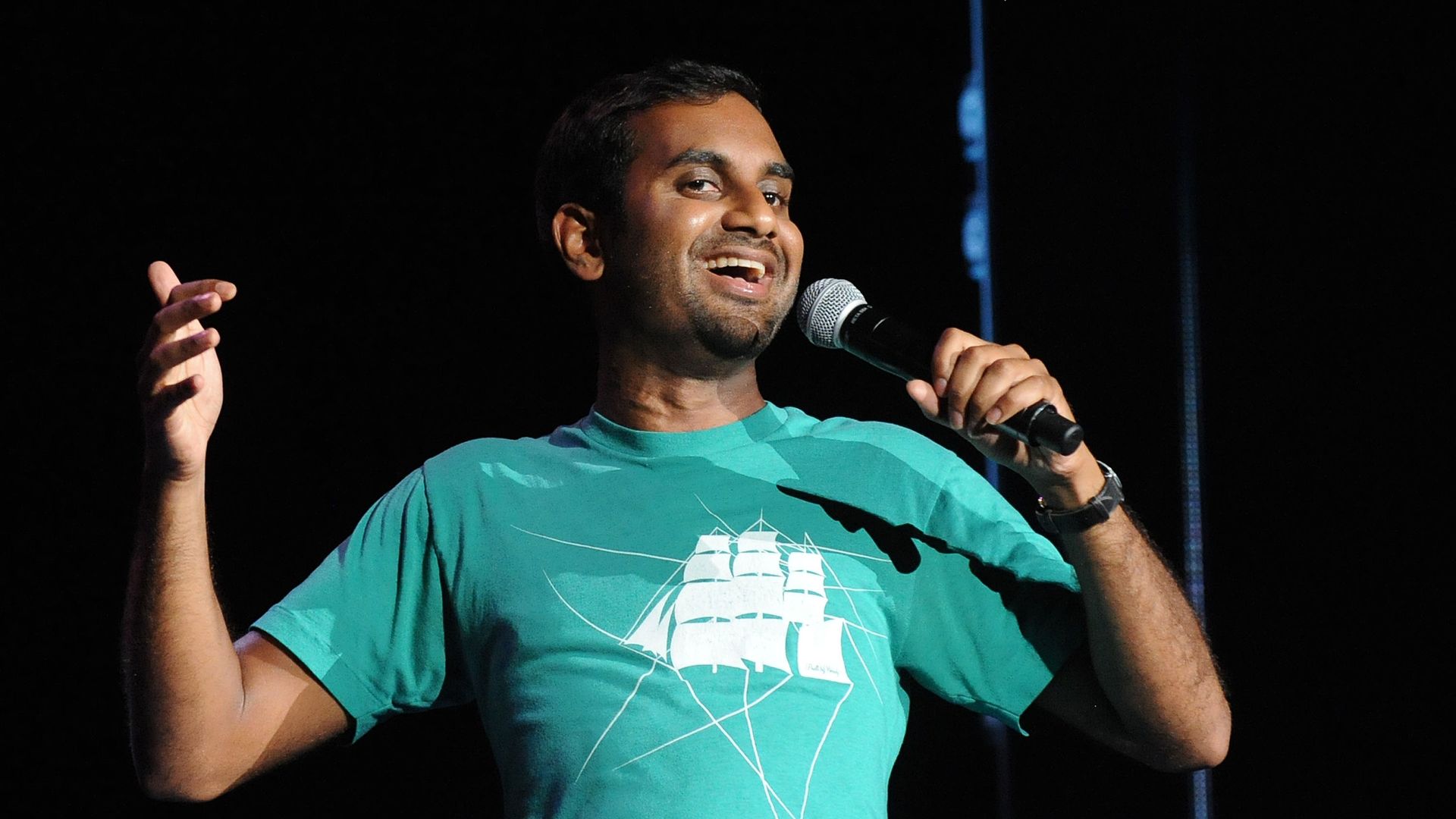 Comedian Aziz Ansari performs standup on stage in a green shirt.
