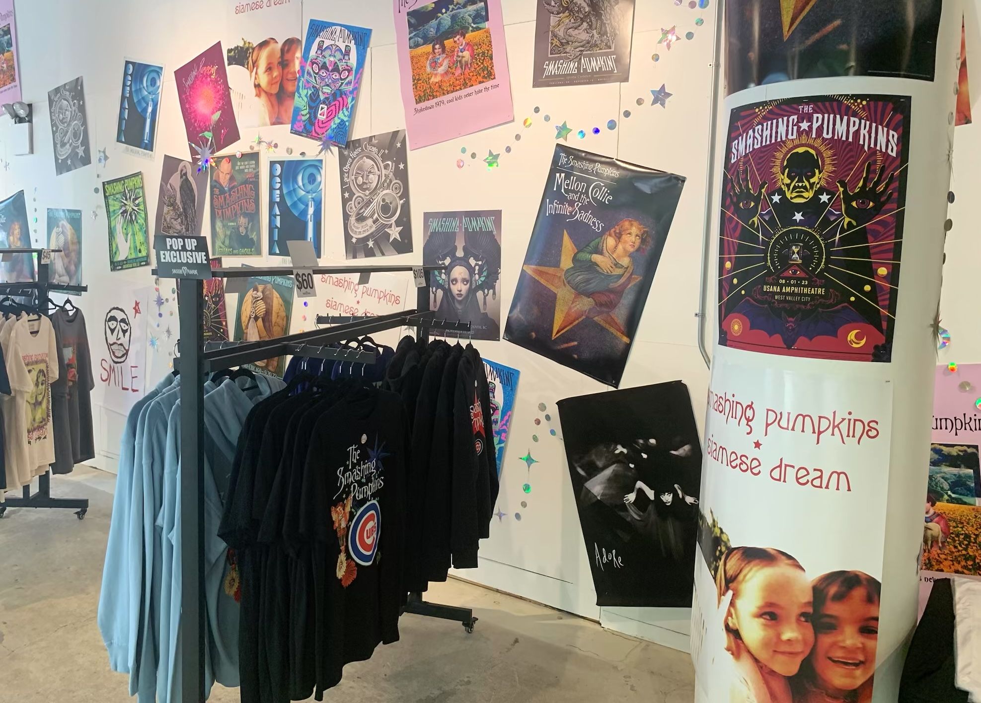 Wall filled with colorful band posters, dominated by "The Smashing Pumpkins", with blue and black shirts hanging below on a clothing rack.