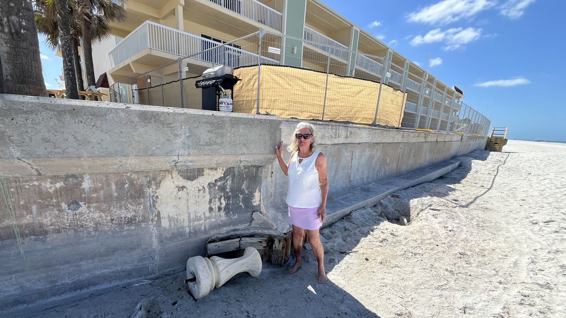 A woman in a white top and lilac skirt stands beside a concrete wall jutting out from sand. A three-story building looms in the background.