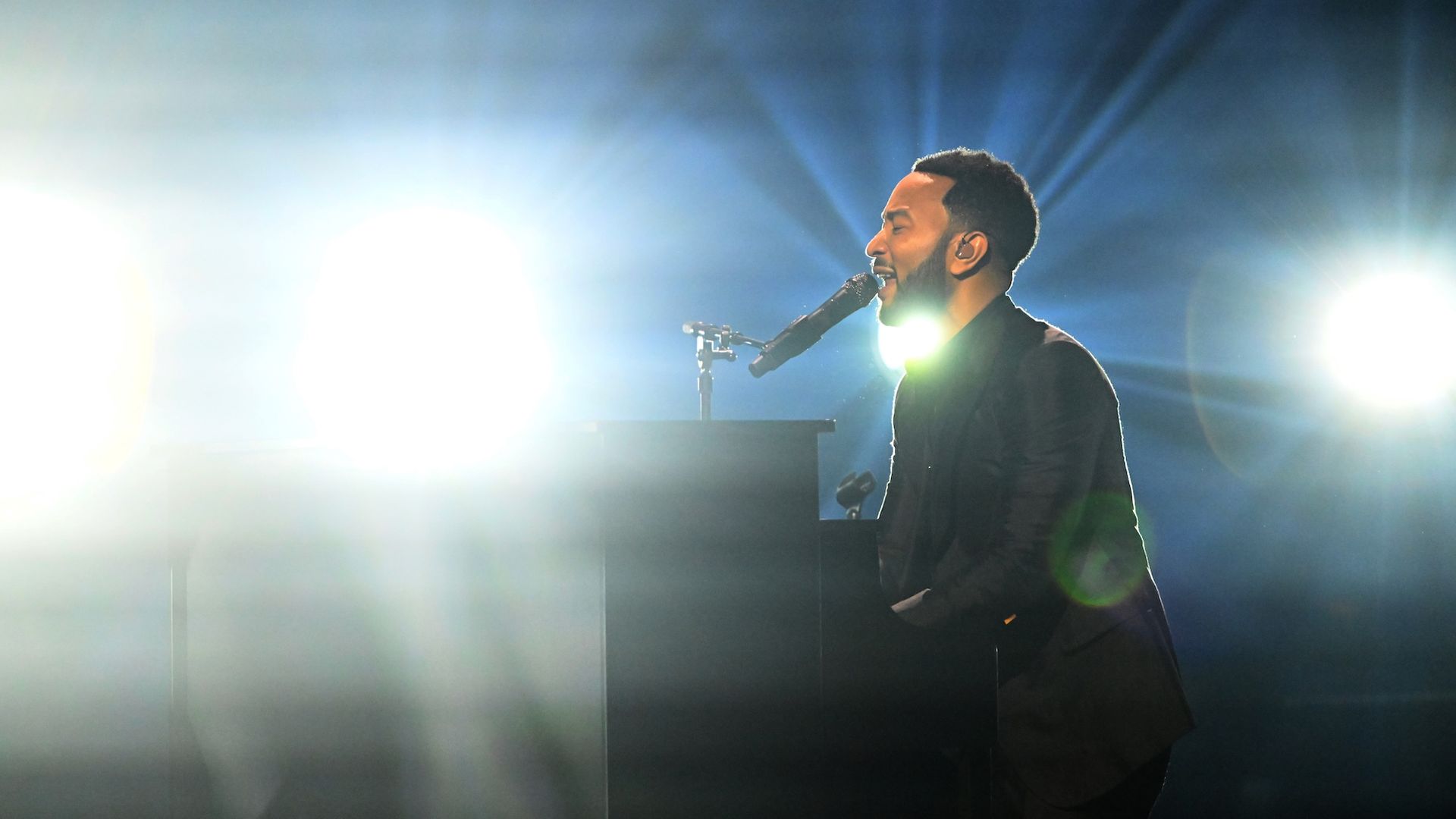 John Legend sings at a piano on stage with bright lights shining behind him.