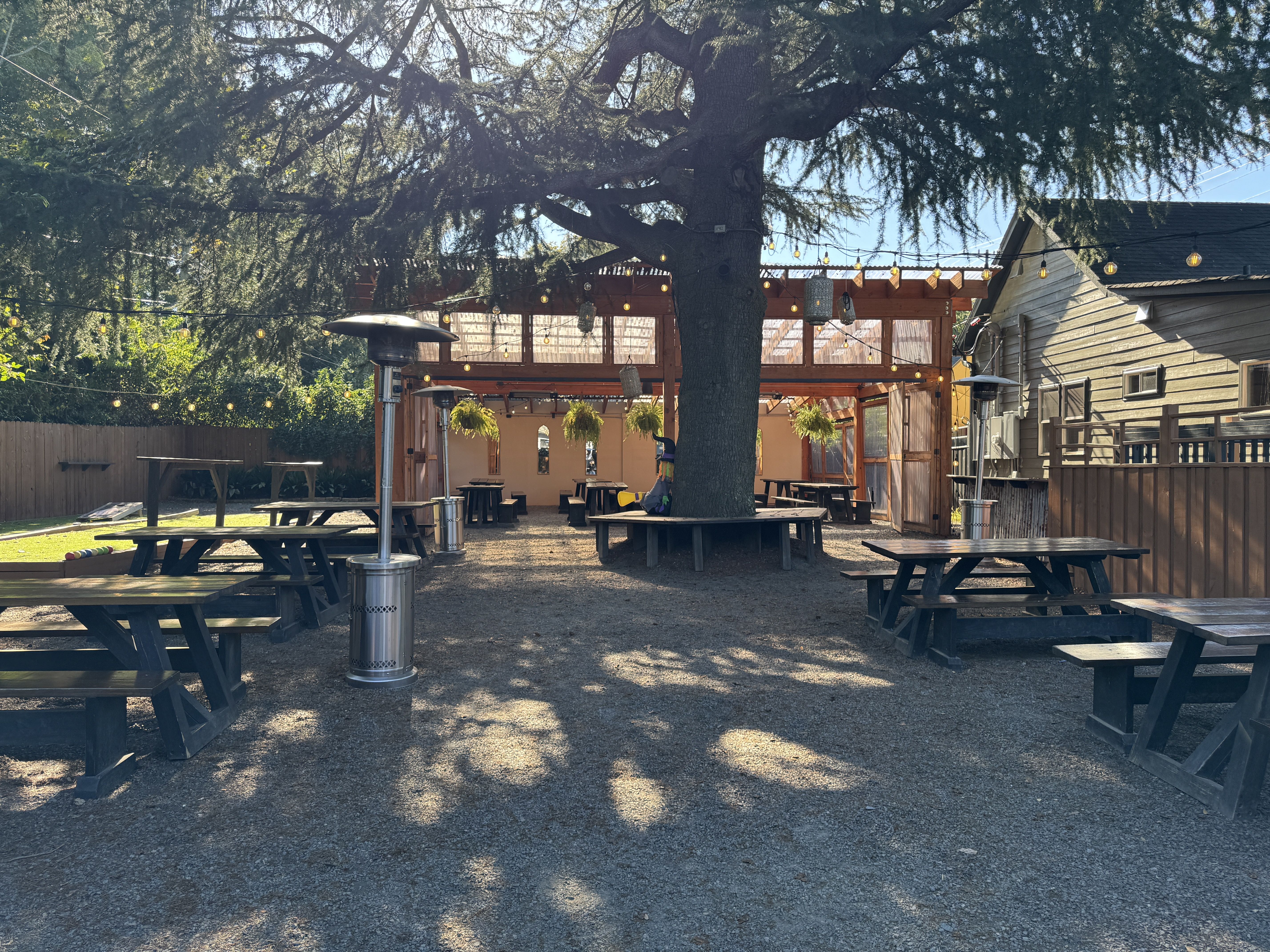 Outdoor patio with a large central tree under a wooden pergola. String lights dangle above rows of picnic tables on a gravel yard, with tall heaters and hanging plants.