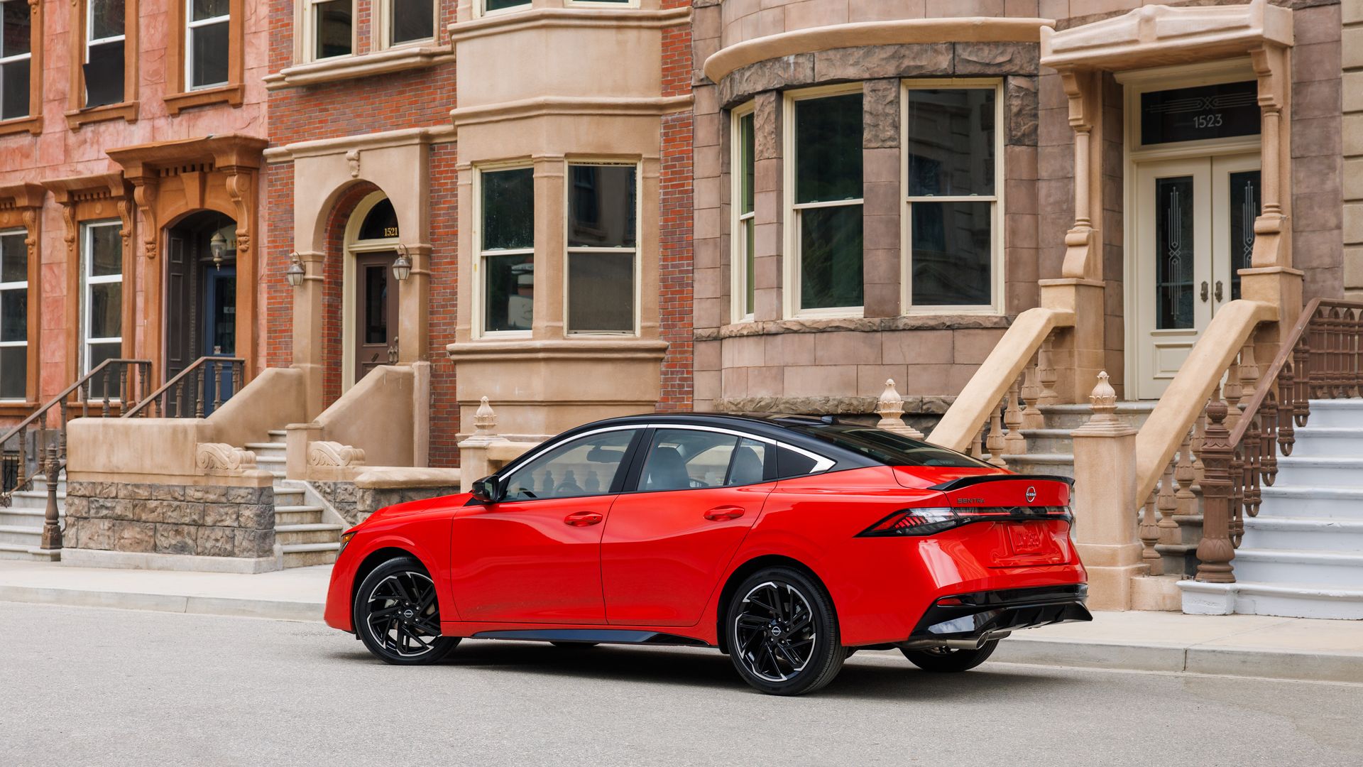 Sporty red Nissan Sentra compact sedan with black roof and wheels parked on a city street in front of brownstone buildings with stairs and large windows.