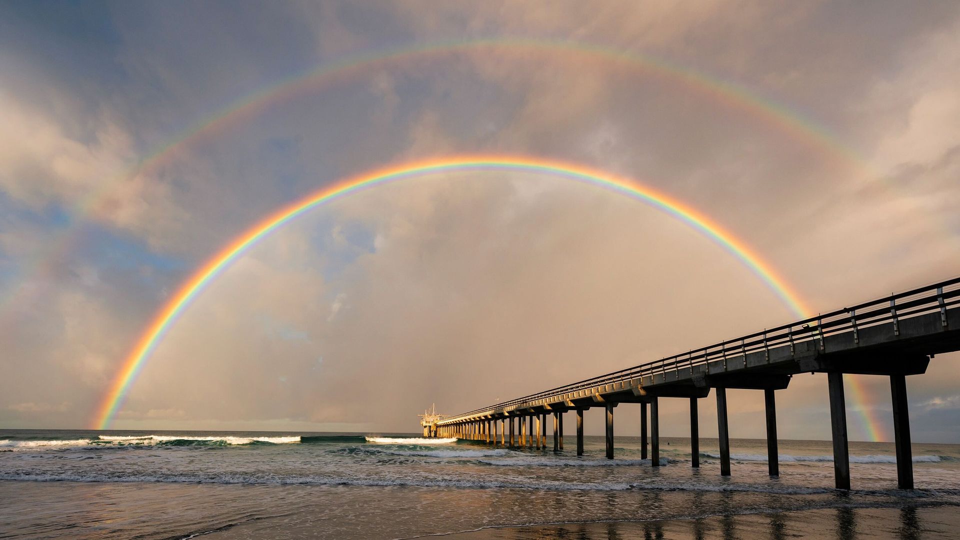 Double rainbow arched over a long pier extending into the ocean with waves and wet sand reflecting the pier and rainbow under a cloudy sky.