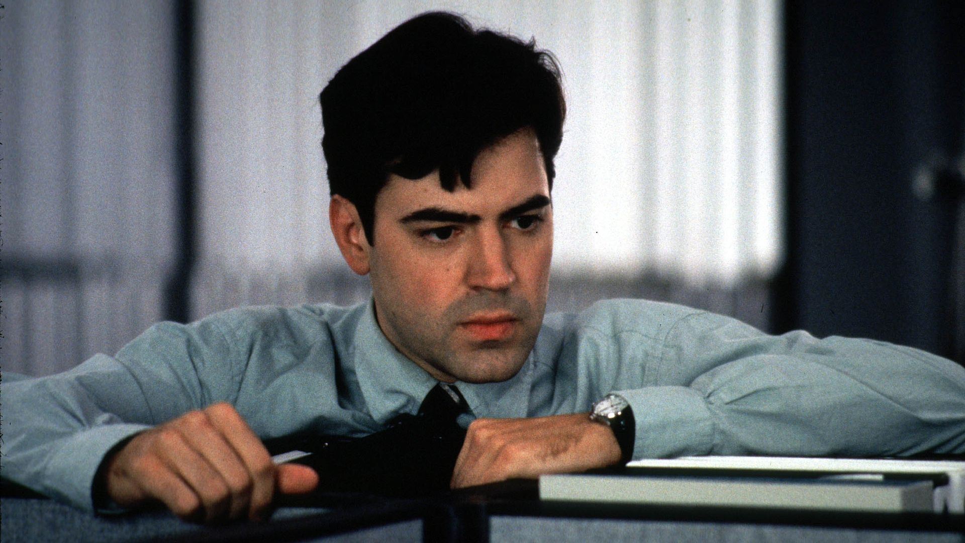 A person with thick black hair, wearing a collared shirt and tie, looks over an office cubicle