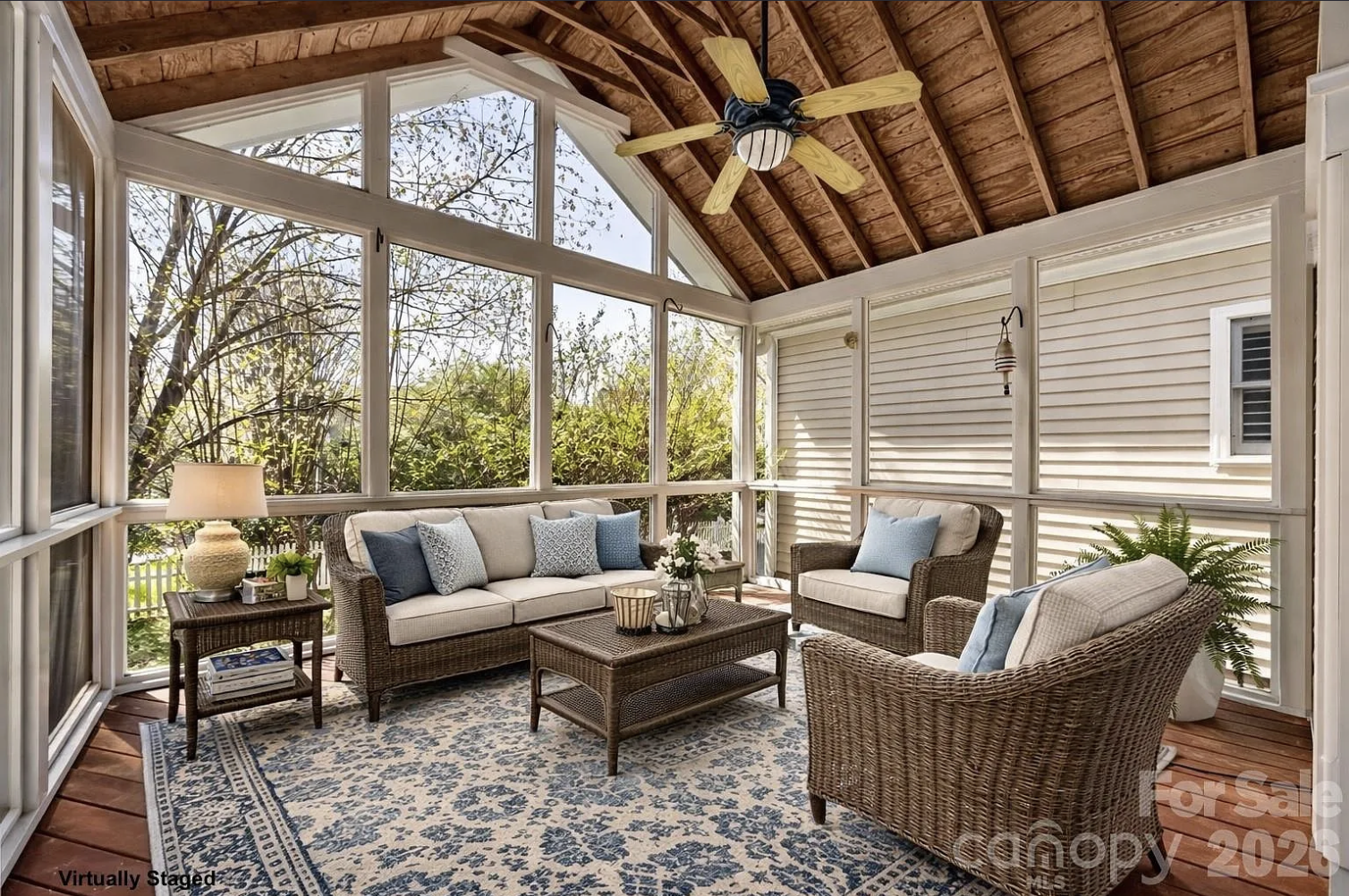 Sunlit screened porch with a vaulted wooden ceiling and large windows, featuring wicker seating: beige sofa and two chairs with blue cushions, a central wicker coffee table, side lamp, and a blue patterned rug.