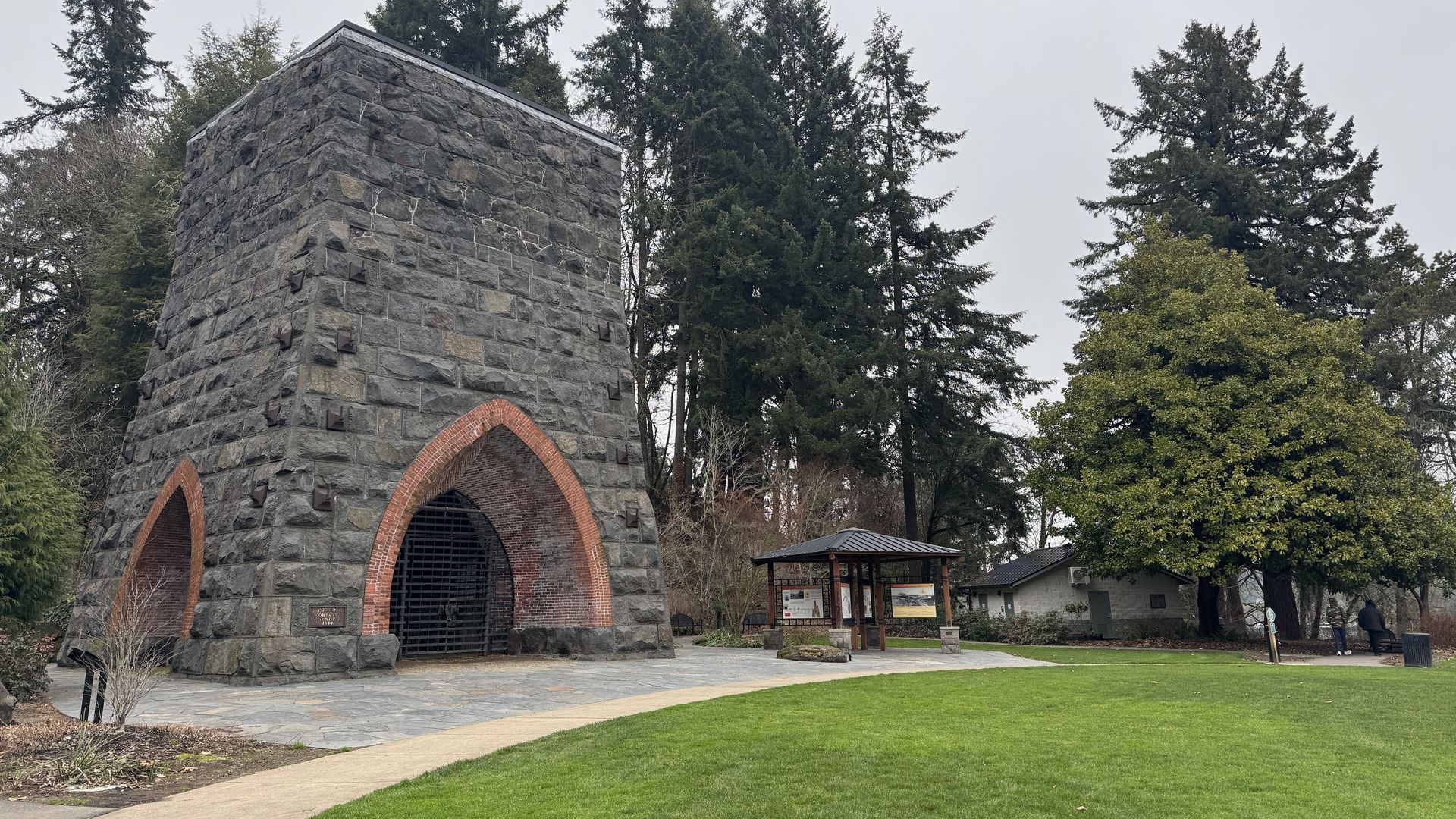 A large stone furnace sits in a park next to a lawn with a small wooden shelter beside it.