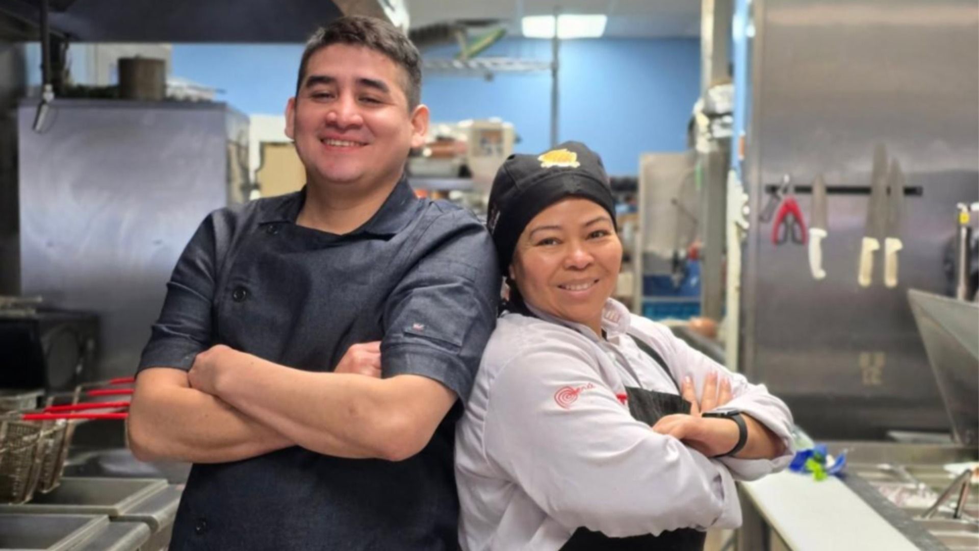 Two smiling chefs with arms crossed, one in black uniform and the other in white with a black headscarf, standing back-to-back in a commercial kitchen with knives on the wall.