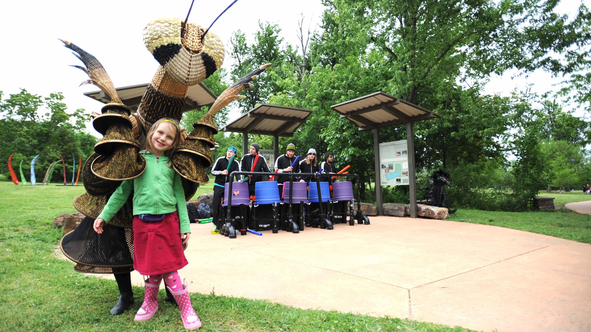A girl stands with a giant woven mantis at a park. 