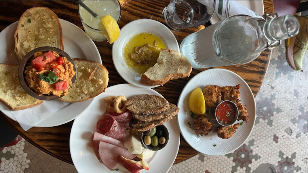 Table with plates of garlic bread with tomato and basil spread, charcuterie with meats, cheese, olives, bite-size snacks with lemon and sauce, a glass with lemon drink, water bottle, and a small plate of bread with olive oil.