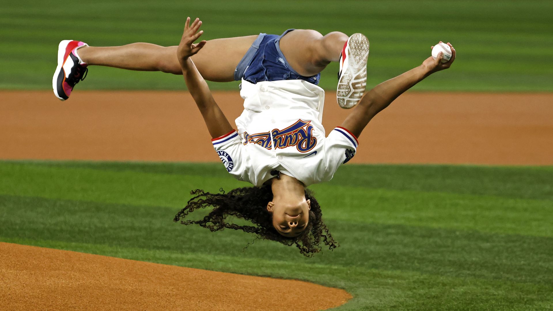 Gymnast Hezly Rivera wearing a Texas Rangers jersey is upside down as she flips to throw a ceremonial first pitch