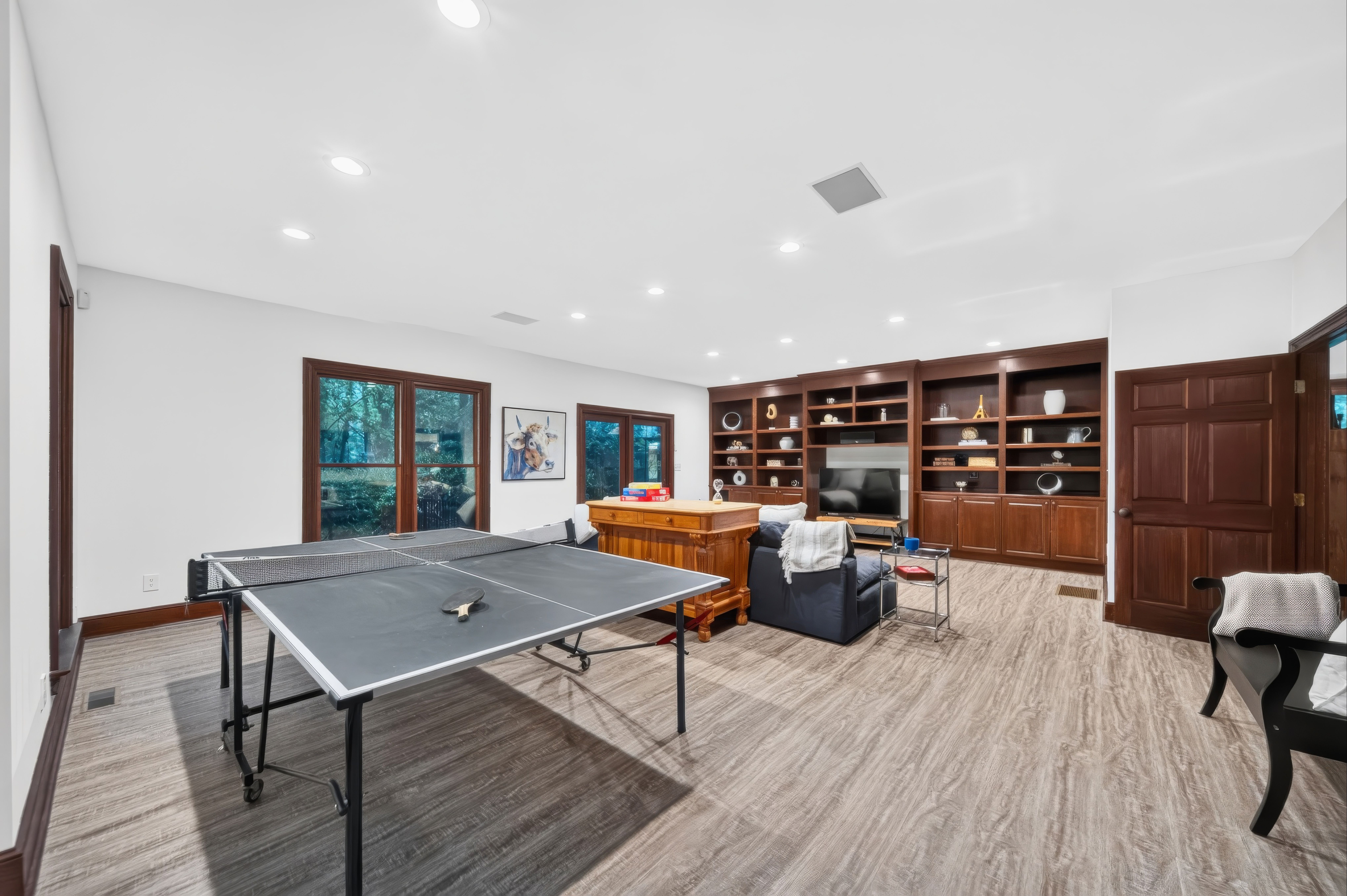 Bright basement room with white ceiling and recessed lights, dark wood built-in shelves and a TV along the back wall. A large table sits in the foreground, and wood-framed windows line the left.