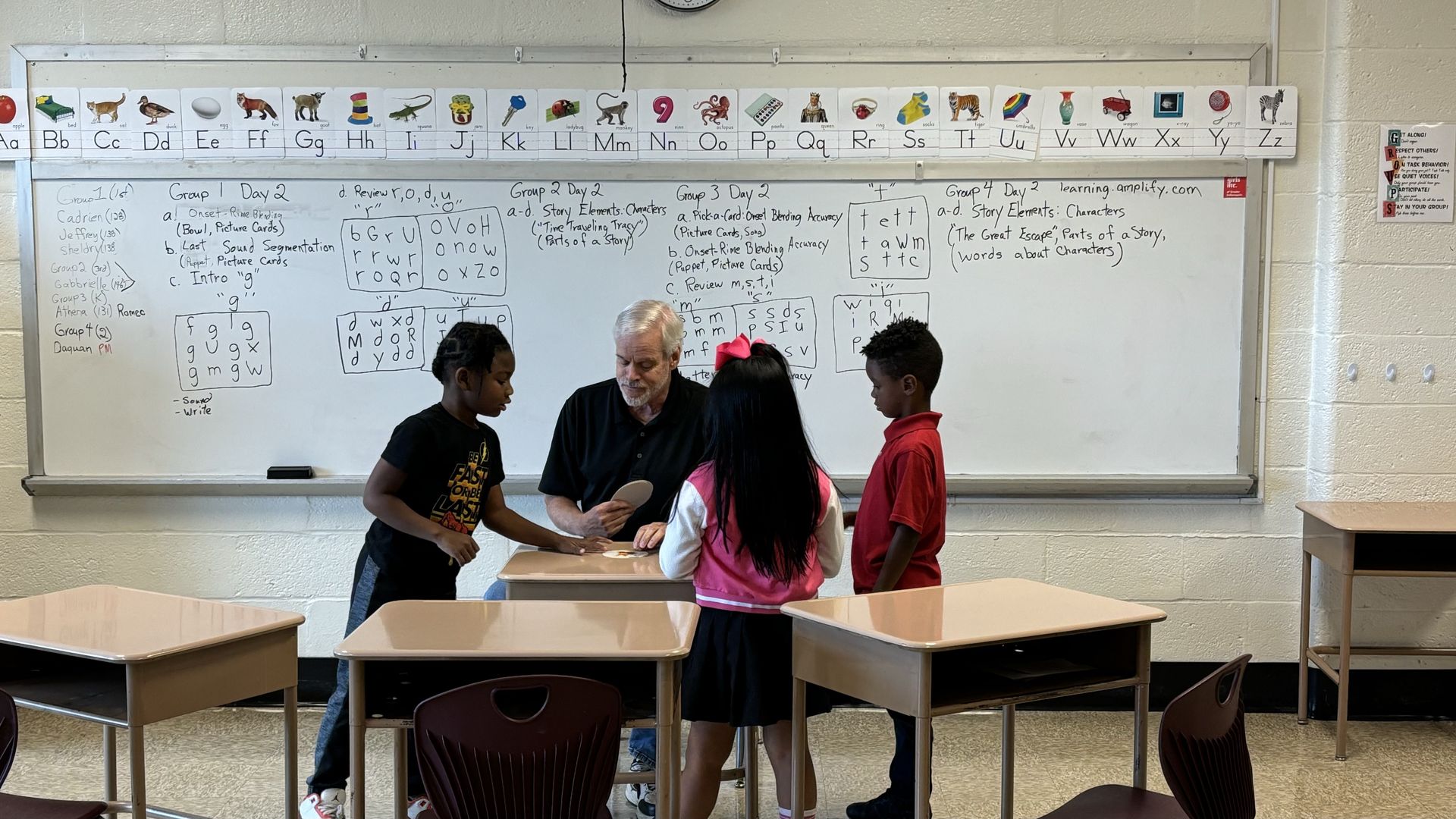 A man sitting at a desk with three kids around him in front of a white board. 