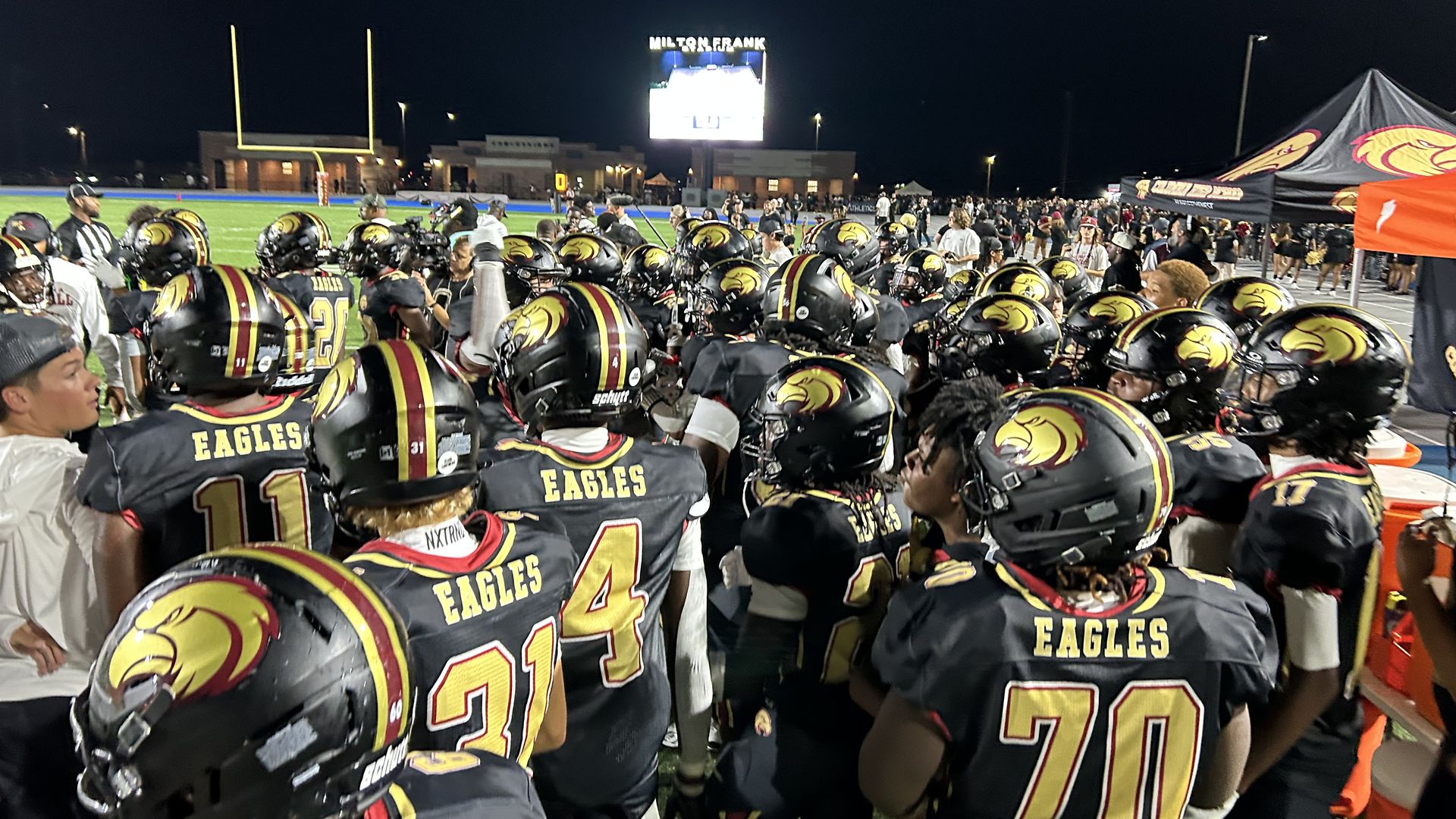 Nighttime football game with players in black and gold Eagles jerseys crowded along the sideline; a bright scoreboard reads Milton Frank, with goalposts, tents, and cheering spectators in the background.