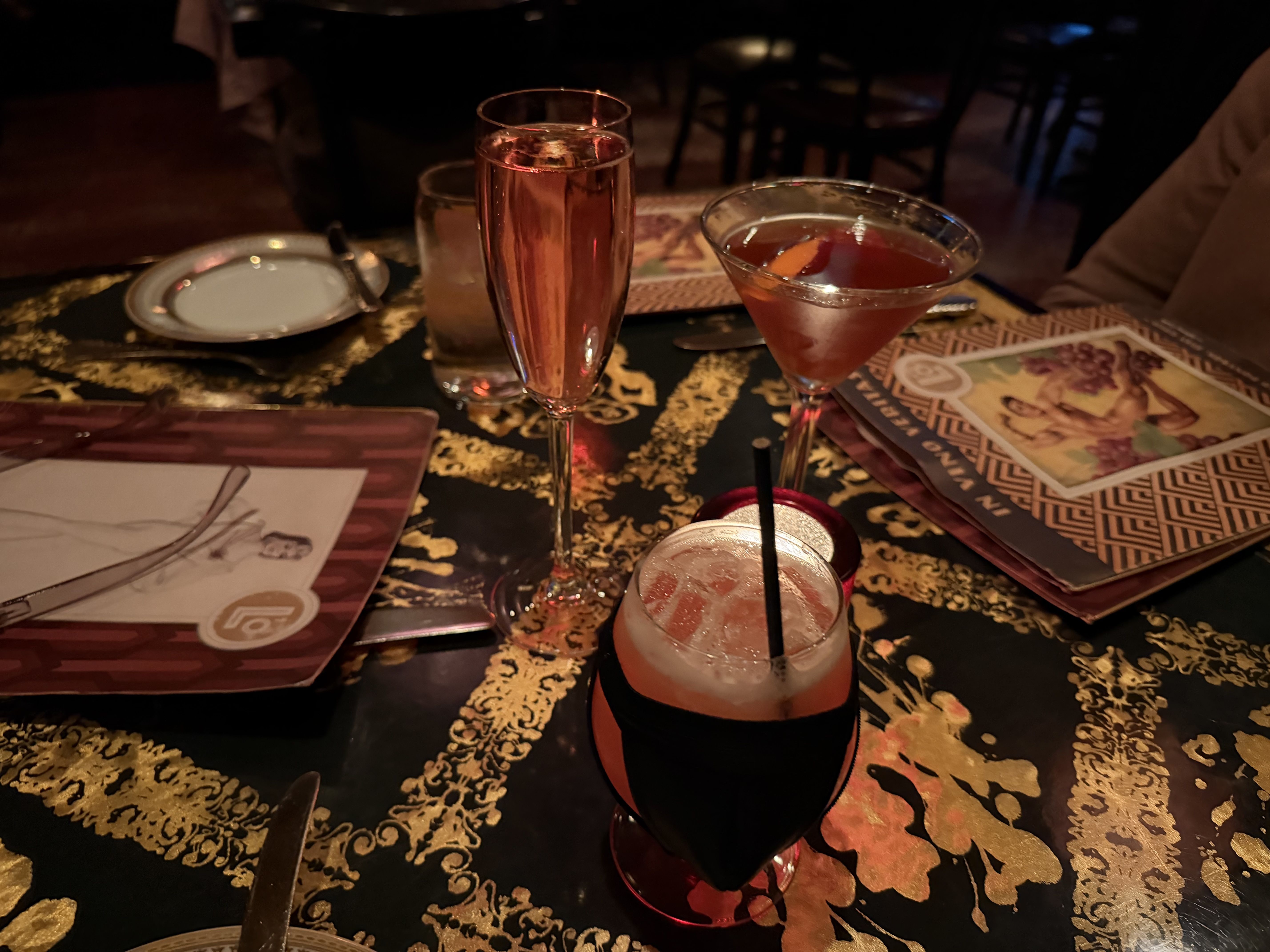 Dimly lit table with three cocktails: a tall pink champagne flute, a red martini glass, and a short frosted drink with a straw. Gold-and-black patterned tablecloth, plates, and menus nearby.