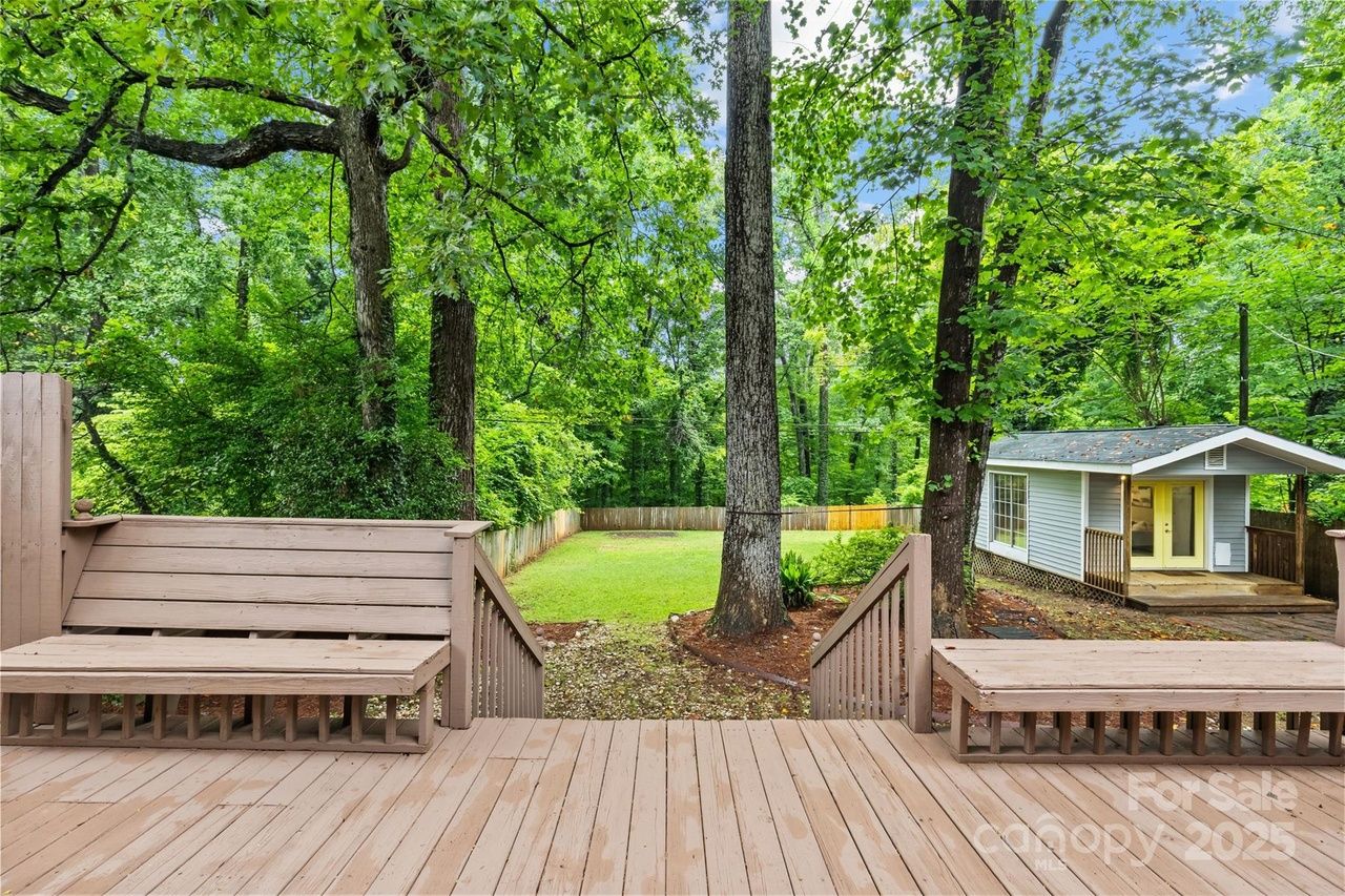 Wooden deck with built-in benches overlooks a fenced green backyard with tall leafy trees and a small grey shed with yellow doors on the right under a blue sky.