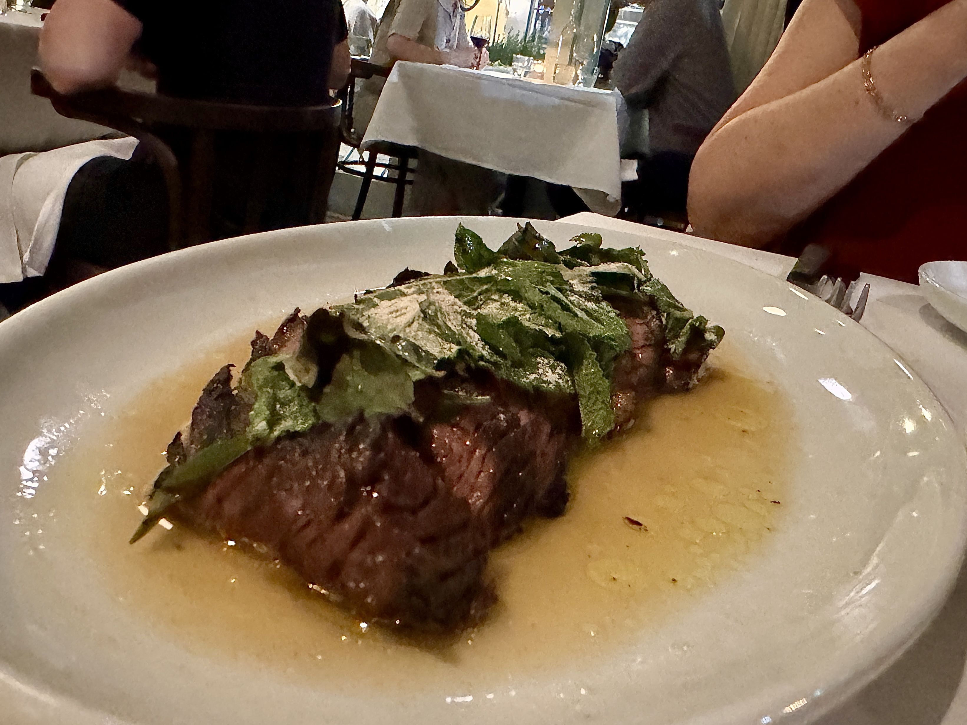 Grilled steak topped with green herbs on a white plate with sauce, served in a restaurant with other diners visible in the background.