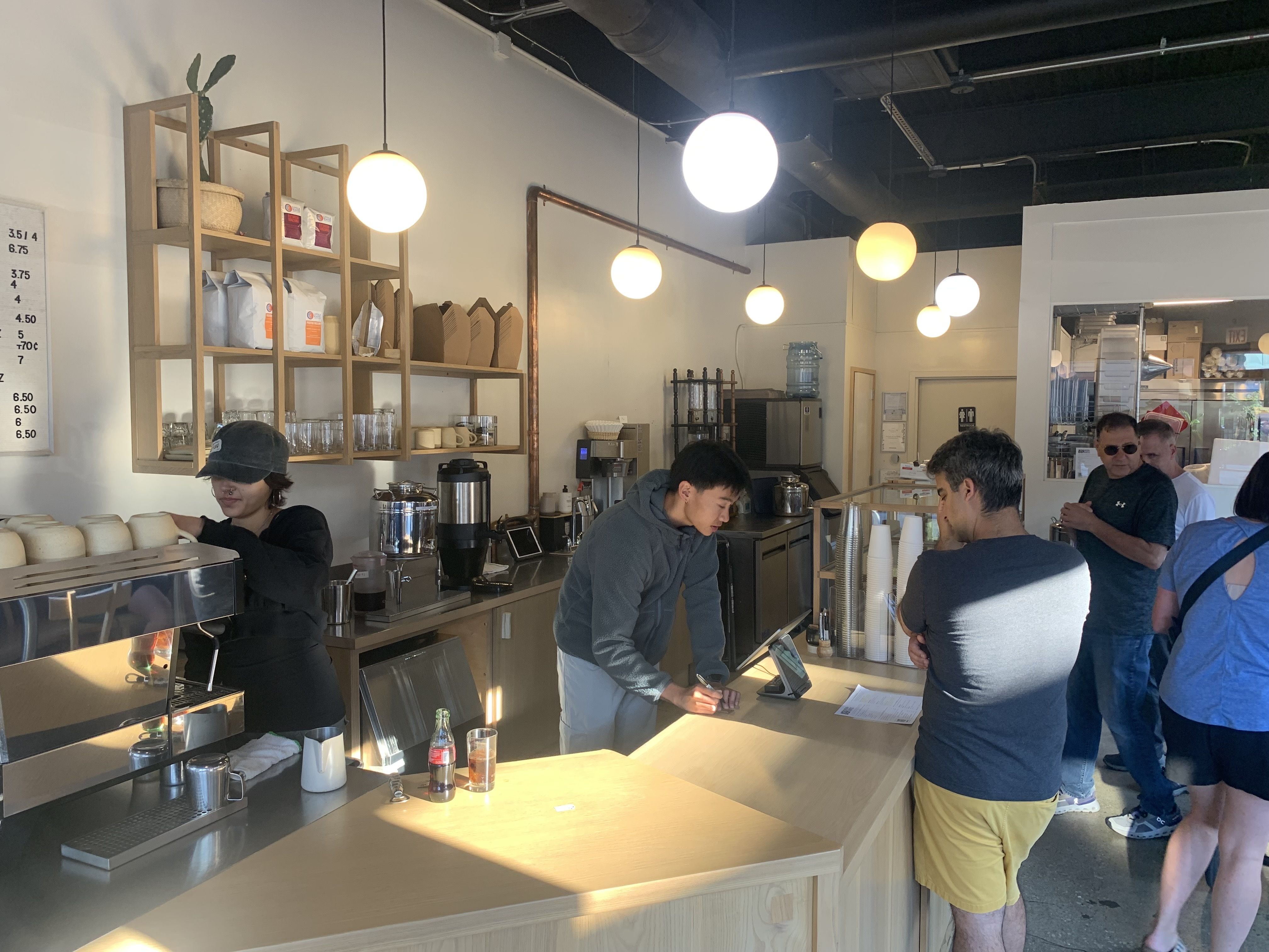 People ordering and working inside a modern cafe with light wooden counters, hanging round lights, shelves with products, and a barista making coffee.