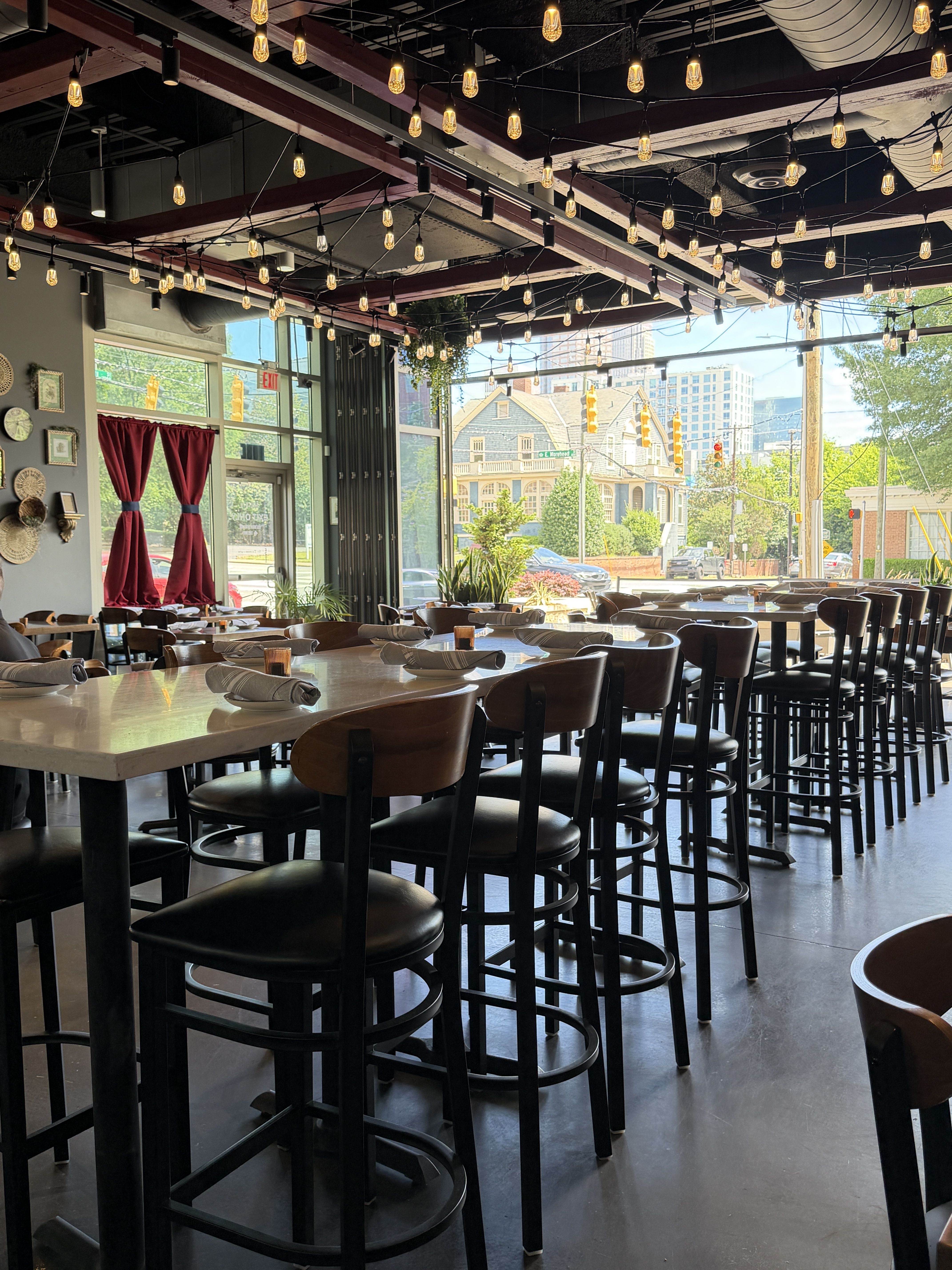 Inside a modern cafe with string lights across a high ceiling, rows of tables with rolled napkins and black stools. Large windows reveal a sunny street with houses, and red curtains on the left.