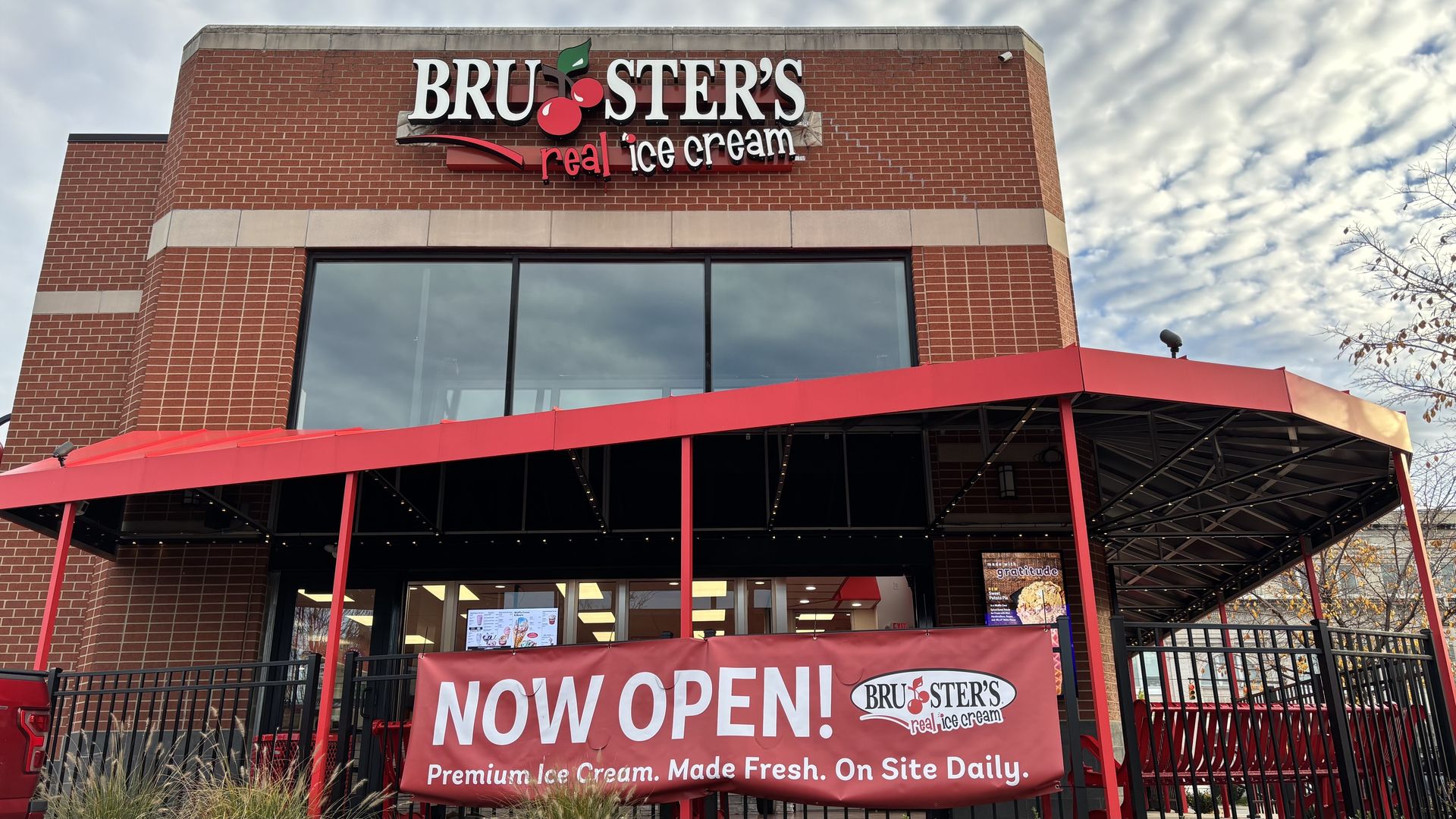 Red brick Bruster's real ice cream shop with large windows, red awning, and red banner reading "NOW OPEN! Premium Ice Cream. Made Fresh. On Site Daily." under cloudy sky.