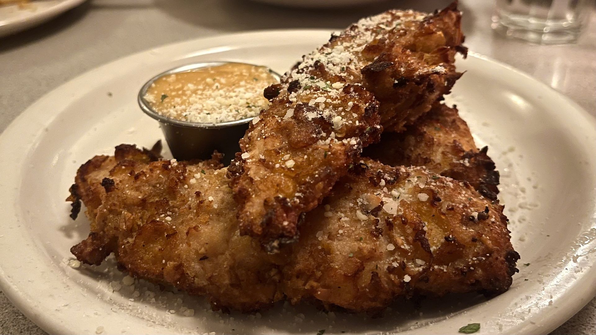 Golden-brown fried chicken tenders sprinkled with grated cheese, served on a white plate with a small cup of creamy orange dipping sauce.