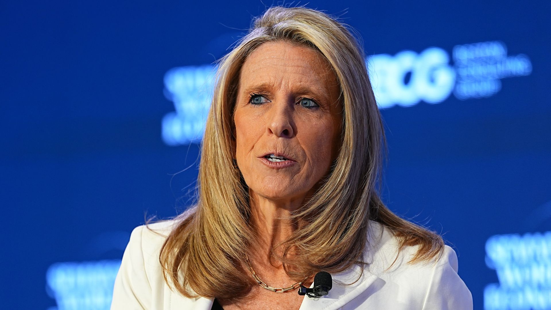Close-up of a blonde person with blue eyes wearing a white blazer and a gold necklace, speaking at a conference with a small microphone clipped to her lapel; blue backdrop.