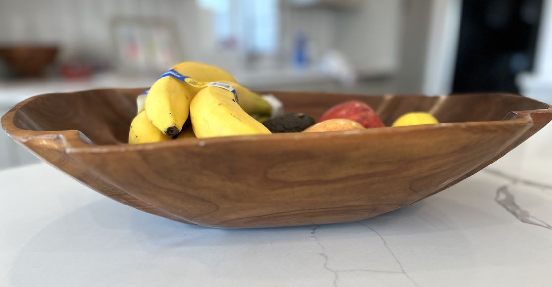 Wooden bowl on a white marble countertop holding bananas, red and yellow apples, an avocado, and a lemon, with a blurred kitchen background.