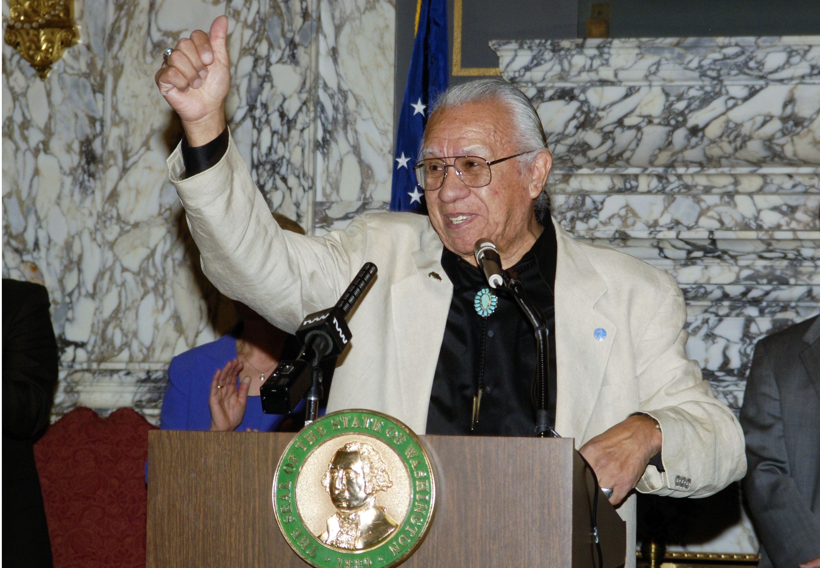 Billy Frank Jr. stands at a podium with the state of Washington seal at the state Capitol, with his right hand raised.