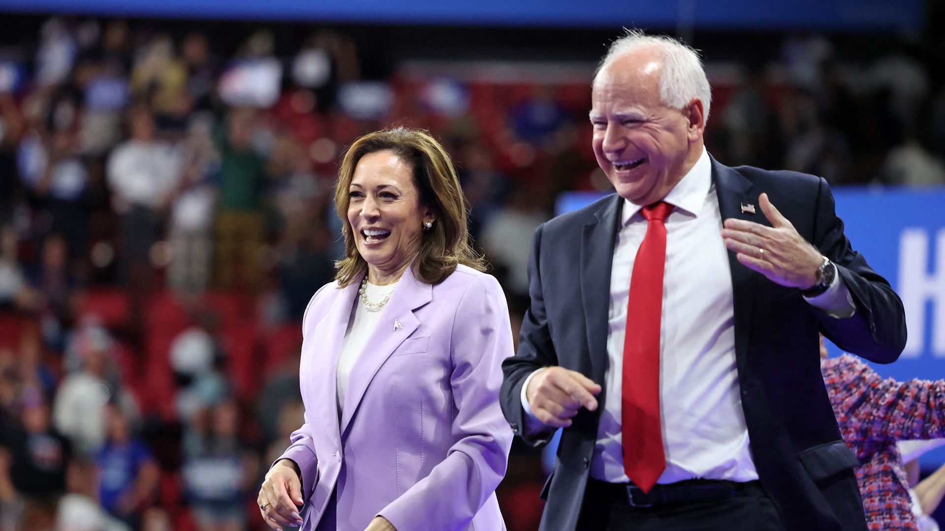 Kamala Harris wearing a light purple blazer and Tim Walz wearing a gray suit stand on stage in front of a large crowd.