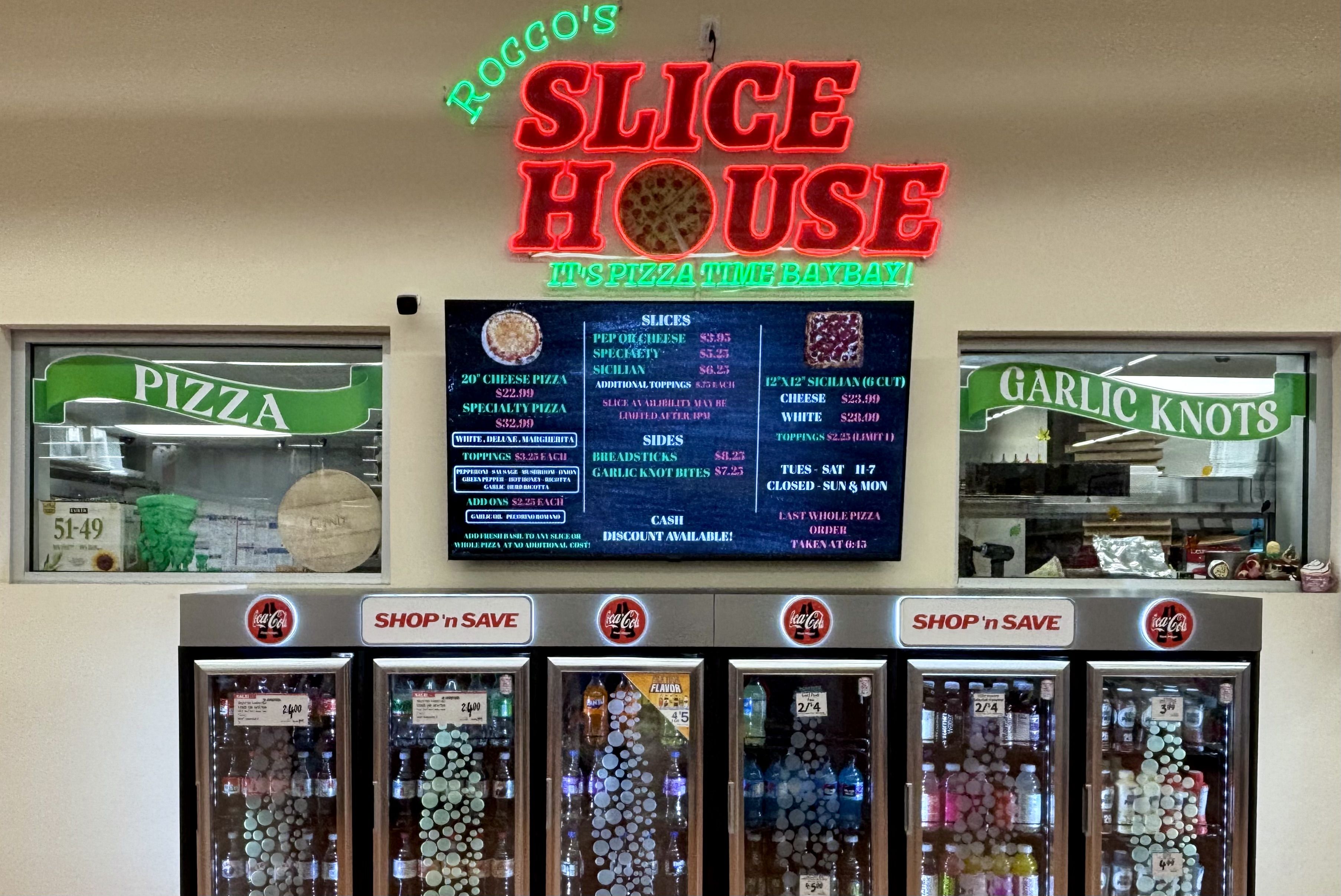 Neon sign says "ROCCO'S SLICE HOUSE" in green and red above a digital pizza menu; storefront with glass-door coolers of drinks and green banners that read "PIZZA" and "GARLIC KNOTS".