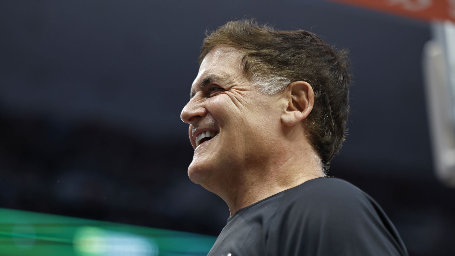Profile of Mark Cuban, a middle-aged man with brown hair and sideburns, smiling, wearing a black Under Armour shirt against a dark background with blurred green and blue lights.