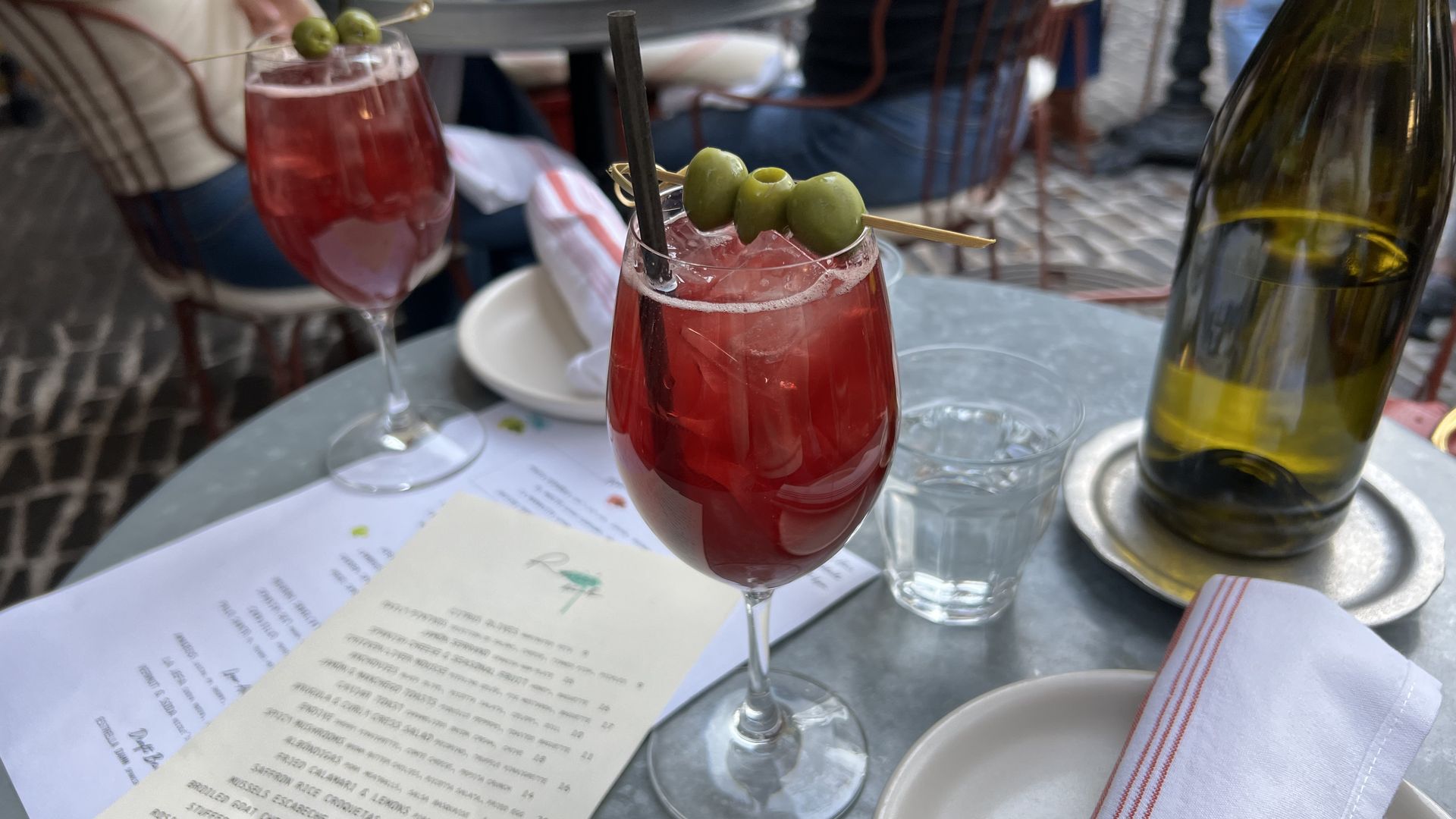 Two red cocktails garnished with green olives on a patio table with menus, a water glass, an empty bottle in a silver holder, and a white plate with a white napkin featuring red stripes.