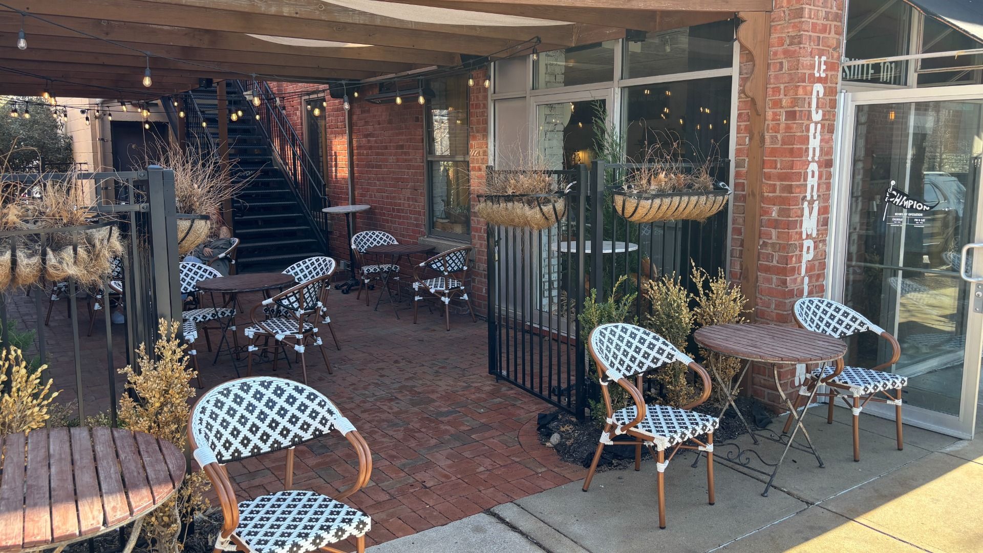 Outdoor cafe patio beside a brick building with string lights and a wooden pergola. Black metal railings, wicker chairs with black-and-white patterned cushions, and round wooden tables; dried grasses in planters.