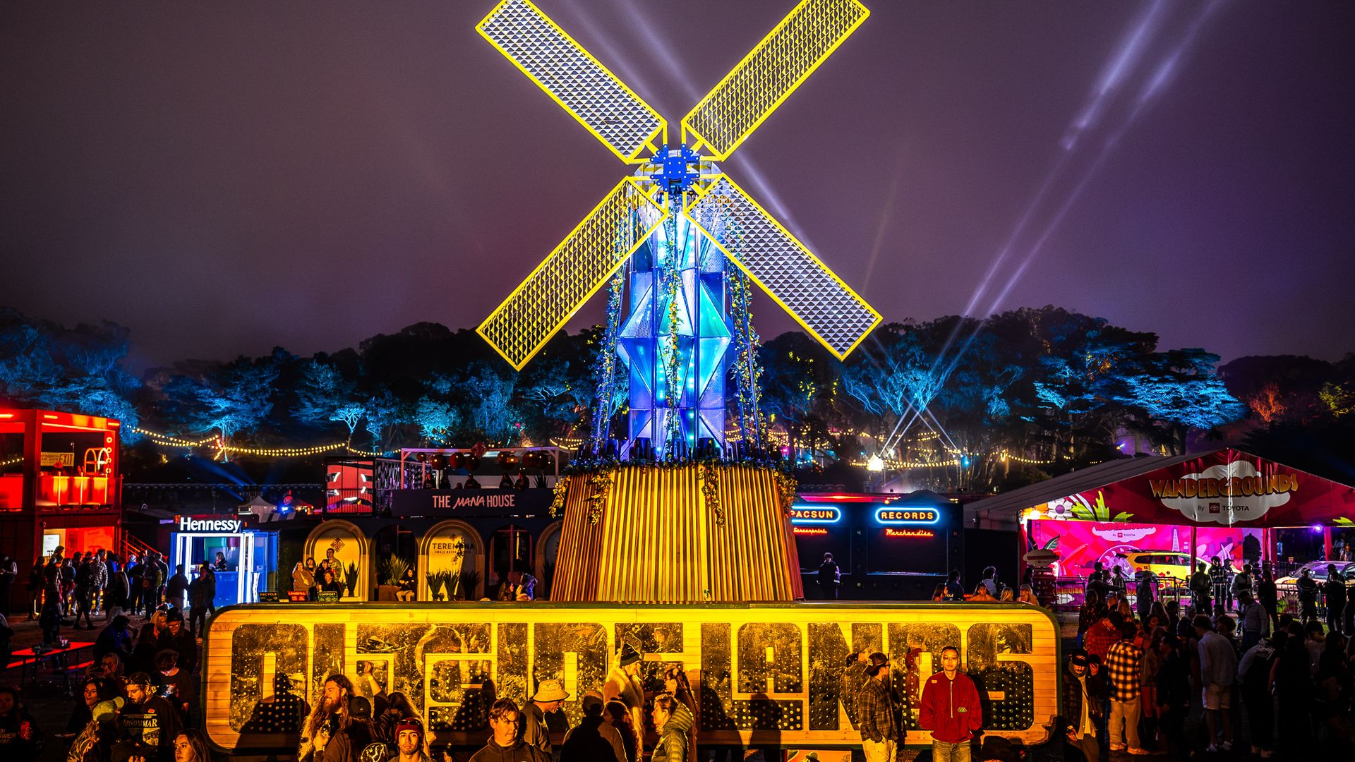 Nighttime festival with a towering neon yellow windmill sculpture at center, blue illuminated base, string lights and vendors, crowd of people in foreground under a purple sky.