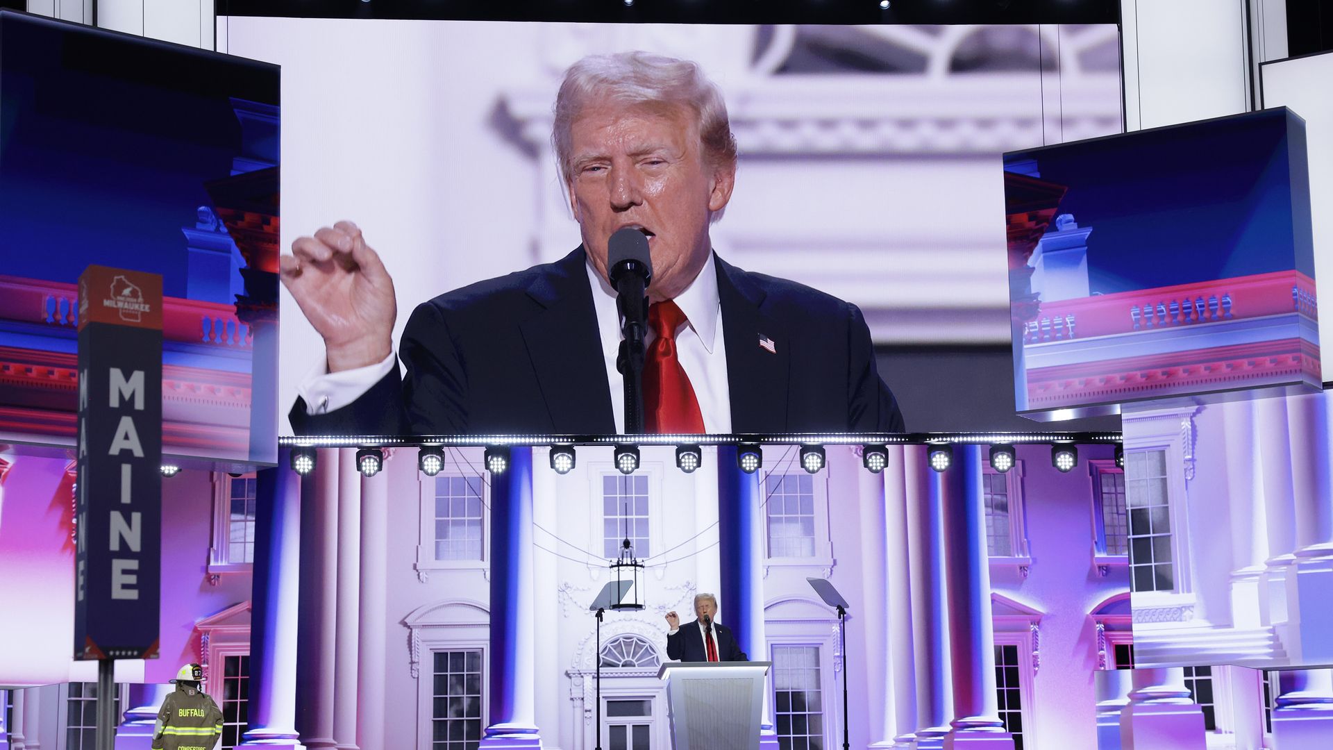 Trump holds his hand up as he speaks from behind a podium on the RNC stage. A large screen displays his movements behind him.