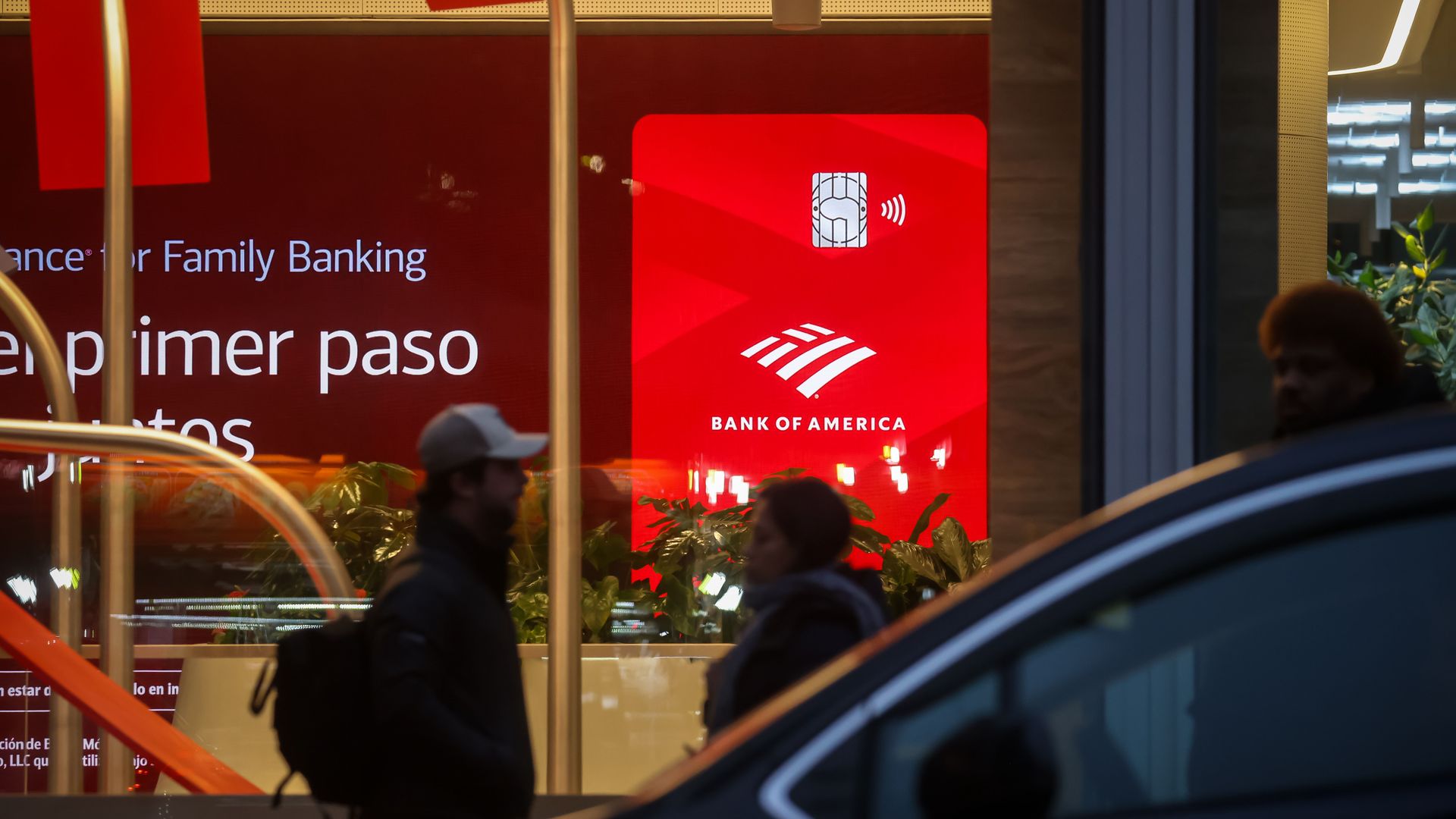 Evening street scene with silhouettes of people walking past a Bank of America branch featuring a large red sign with its logo and text in Spanish about family banking.