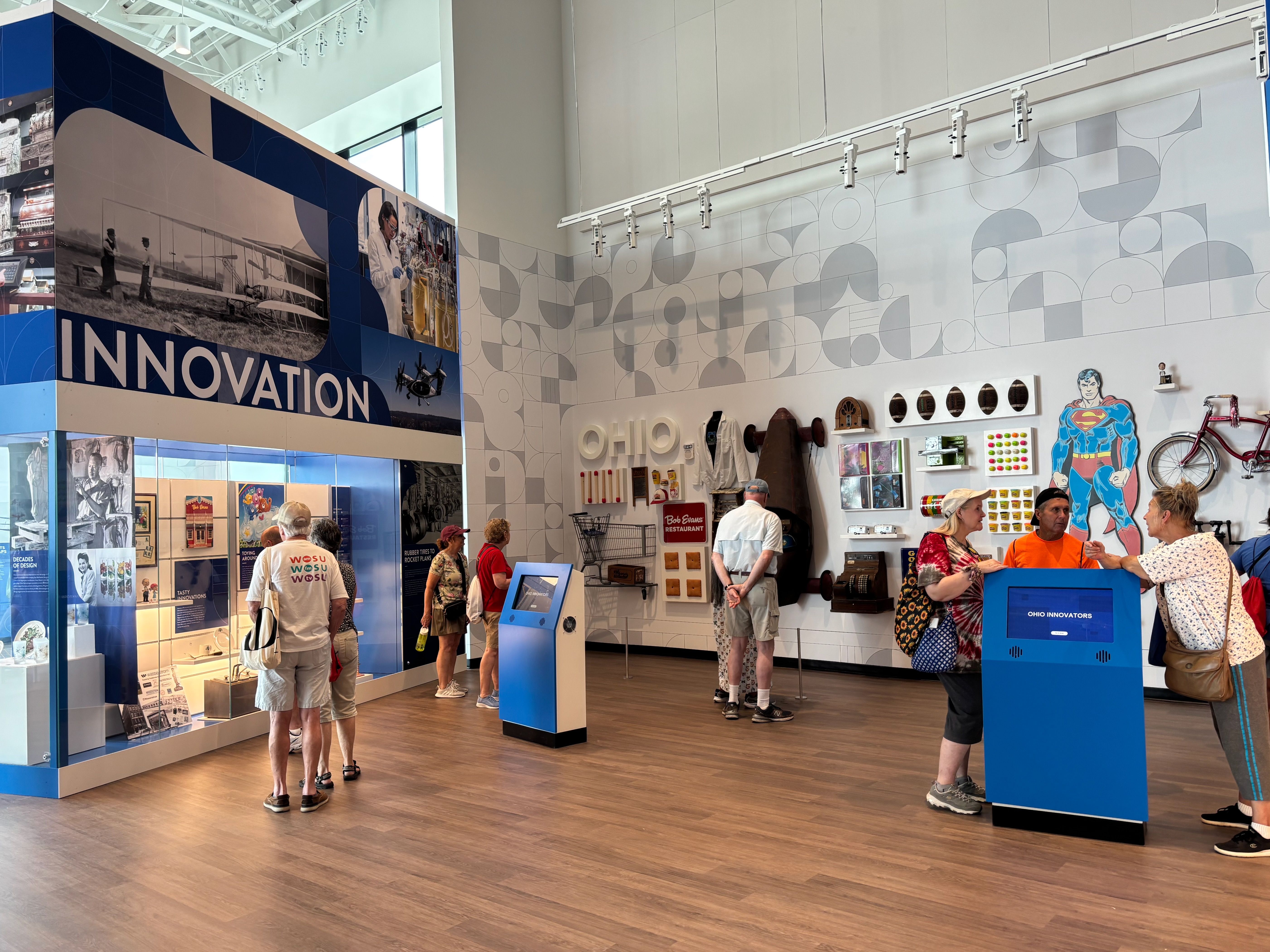 Indoor science-innovation exhibit with blue and gray geometric walls, a large "INNOVATION" sign, visitors browsing displays, a Superman cutout on the right, and shelves of objects.