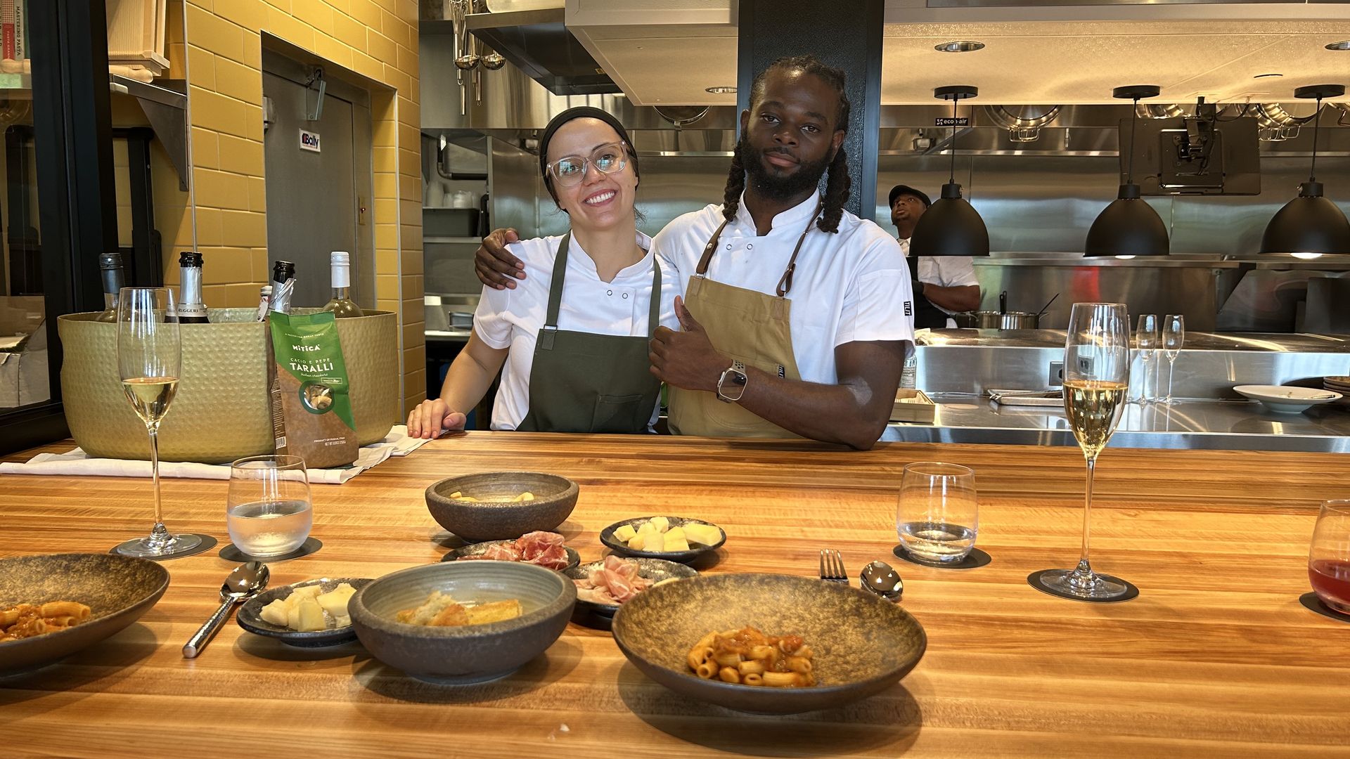 Two chefs in white shirts and aprons smile behind a wooden counter with pasta dishes, cheese, and drinks. Kitchen with yellow tiles and stainless steel equipment in background.