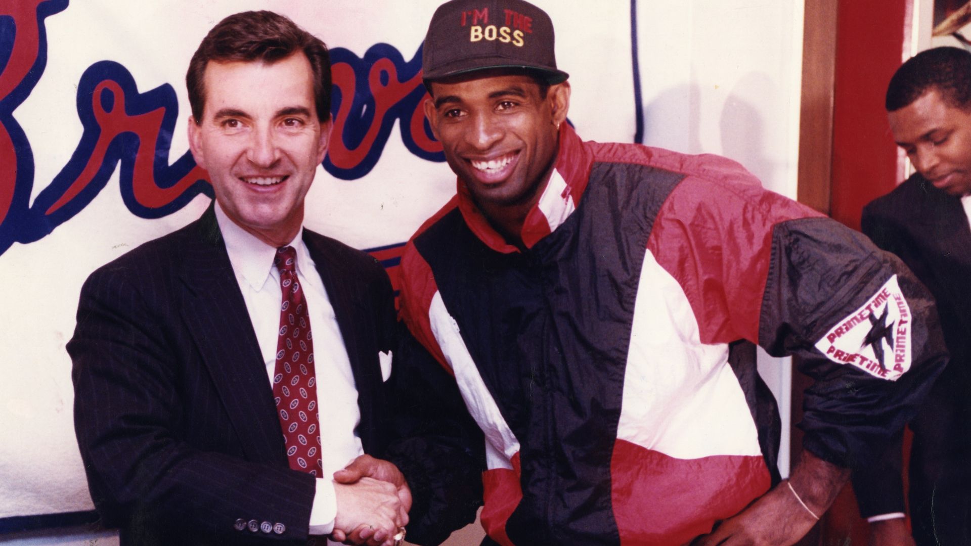 A man in a suit shakes hands in front of an Atlanta Braves sign with an athlete wearing a red, black and white track suit and hat reading "I'm the boss"