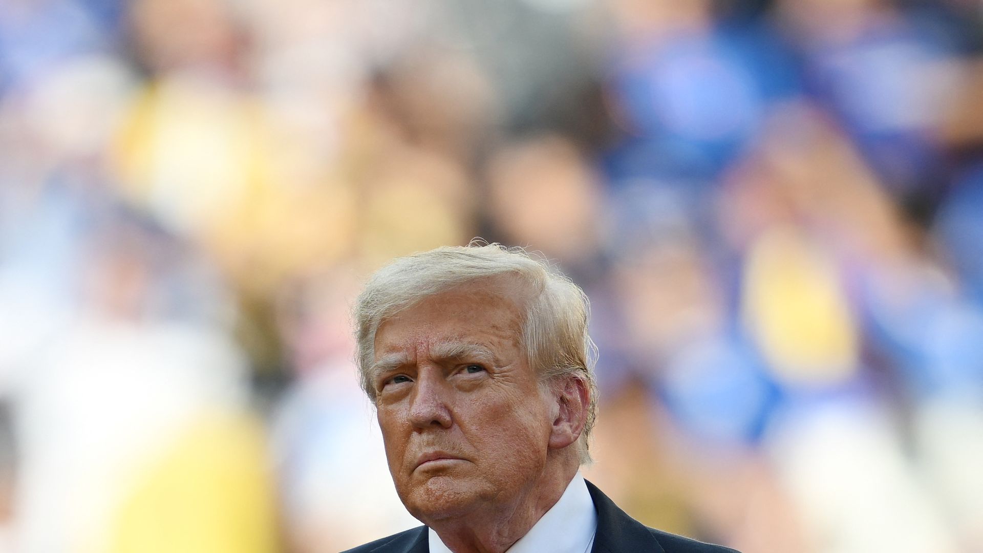 Close-up of Donald Trump wearing a dark suit, white shirt, and red tie, with an American flag pin on his lapel, against a blurred colorful background.