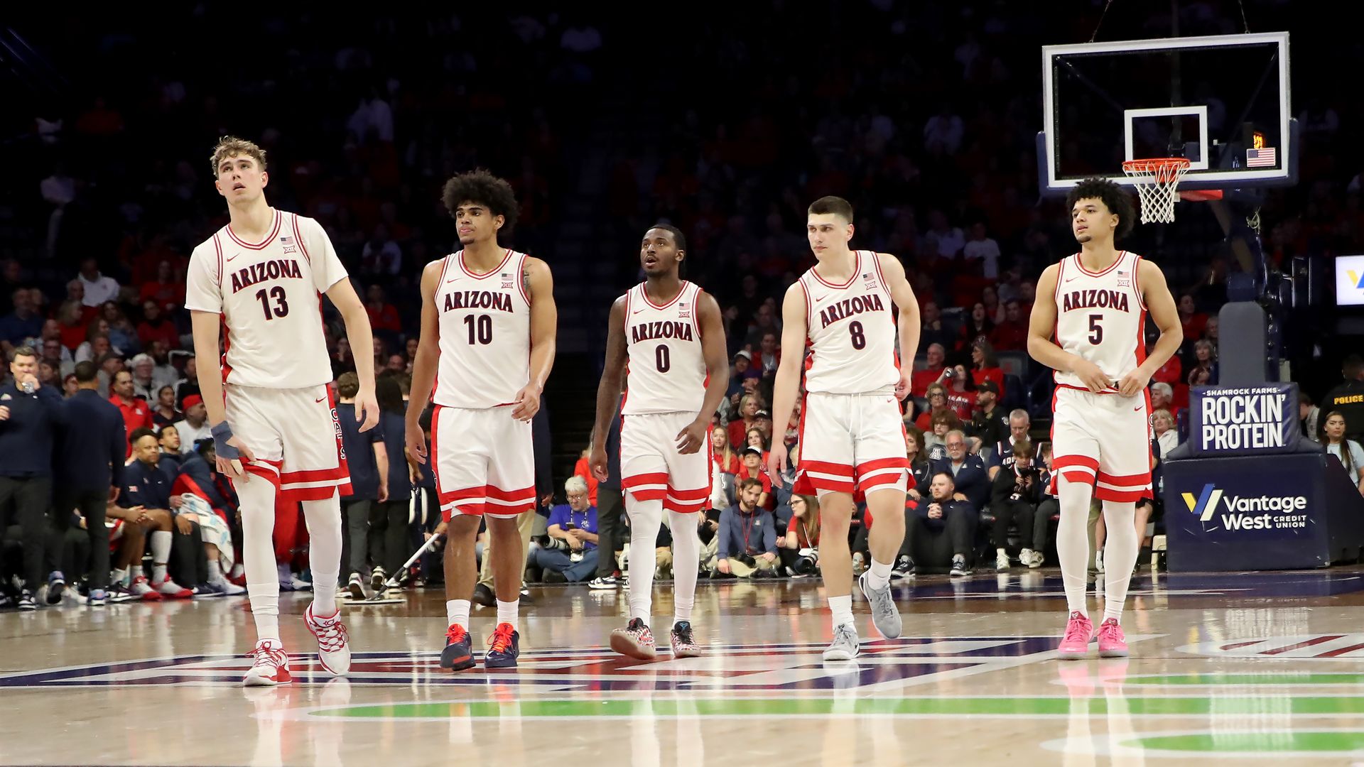 Five basketball players in white uniforms with red trim on the bottom of their shorts, with the word "Arizona" on the front of the shirts, standing on a basketball court with a crowd in the background. 