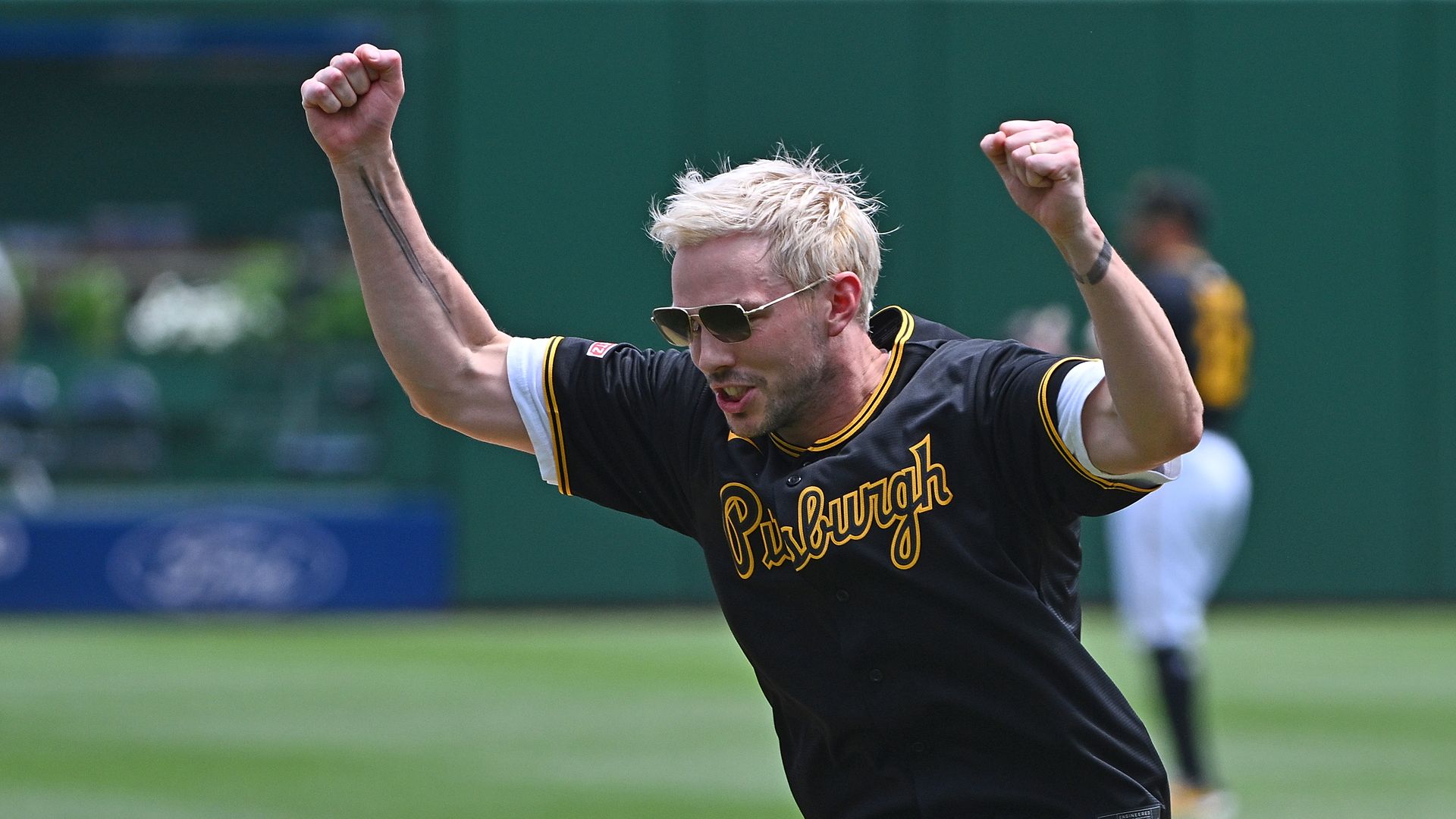 Actor Nicholas Hoult with blonde hair and sunglasses wearing a black Pittsburgh jersey celebrating with fists raised on a baseball field with green backdrop.