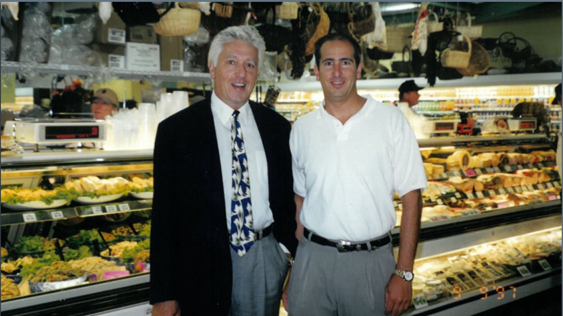 Two men standing and smiling inside a deli or market. One wears a black jacket and patterned tie, the other a white polo shirt. Food displays and hanging baskets in background.