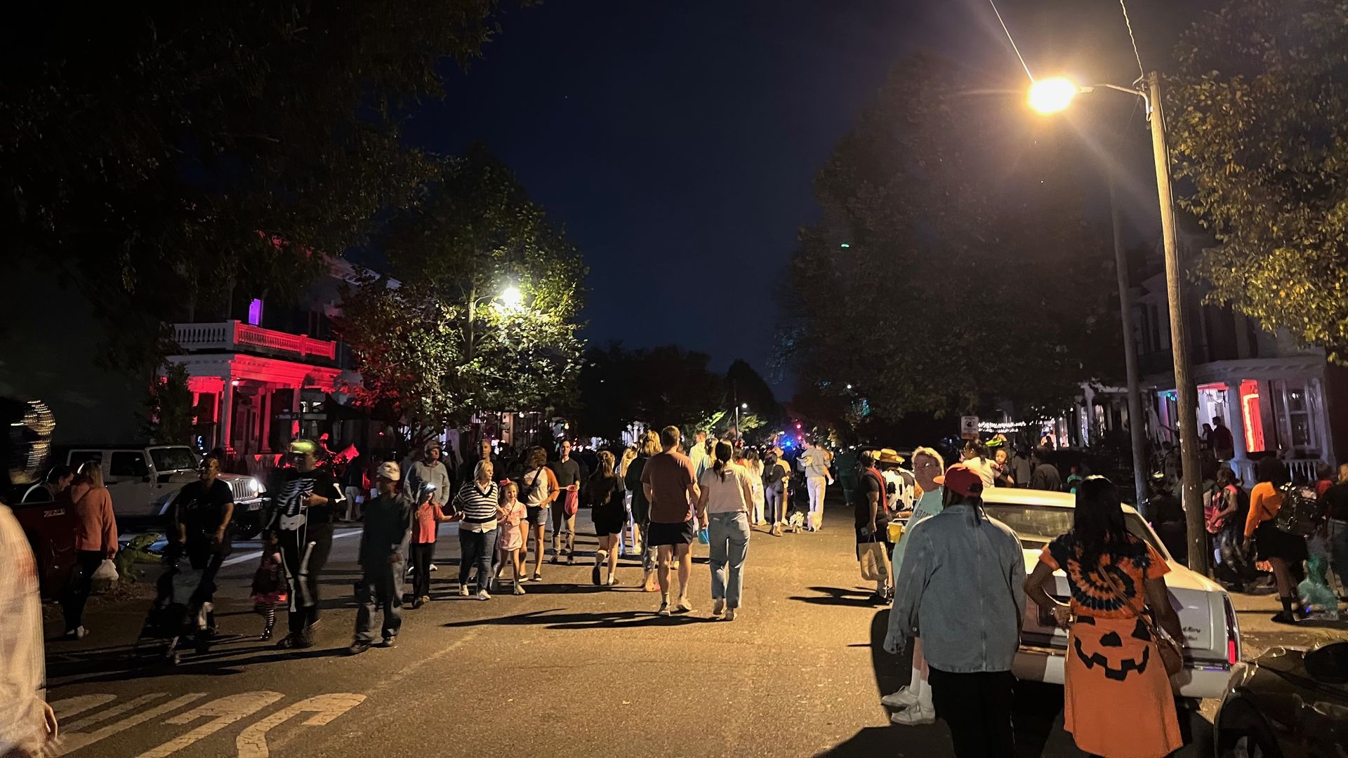 Nighttime street scene with a crowd strolling down a lit avenue. A bright streetlamp on the right glows; red-lit building left, trees frame the scene; a person in a pumpkin costume in the foreground.