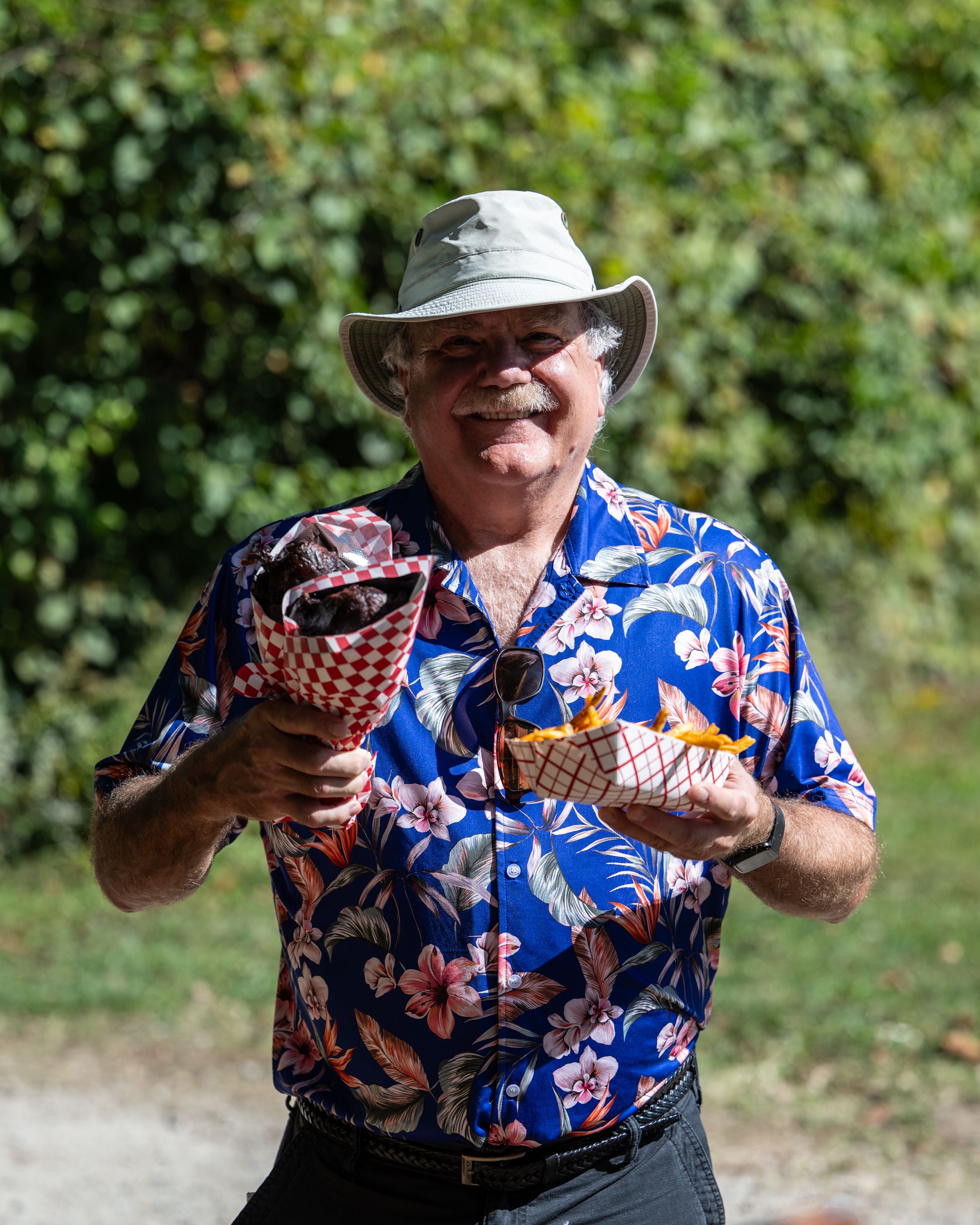 Smiling older man in a beige bucket hat and blue floral Hawaiian shirt holds two red-checkered paper cones of food outdoors, with green foliage in the background.